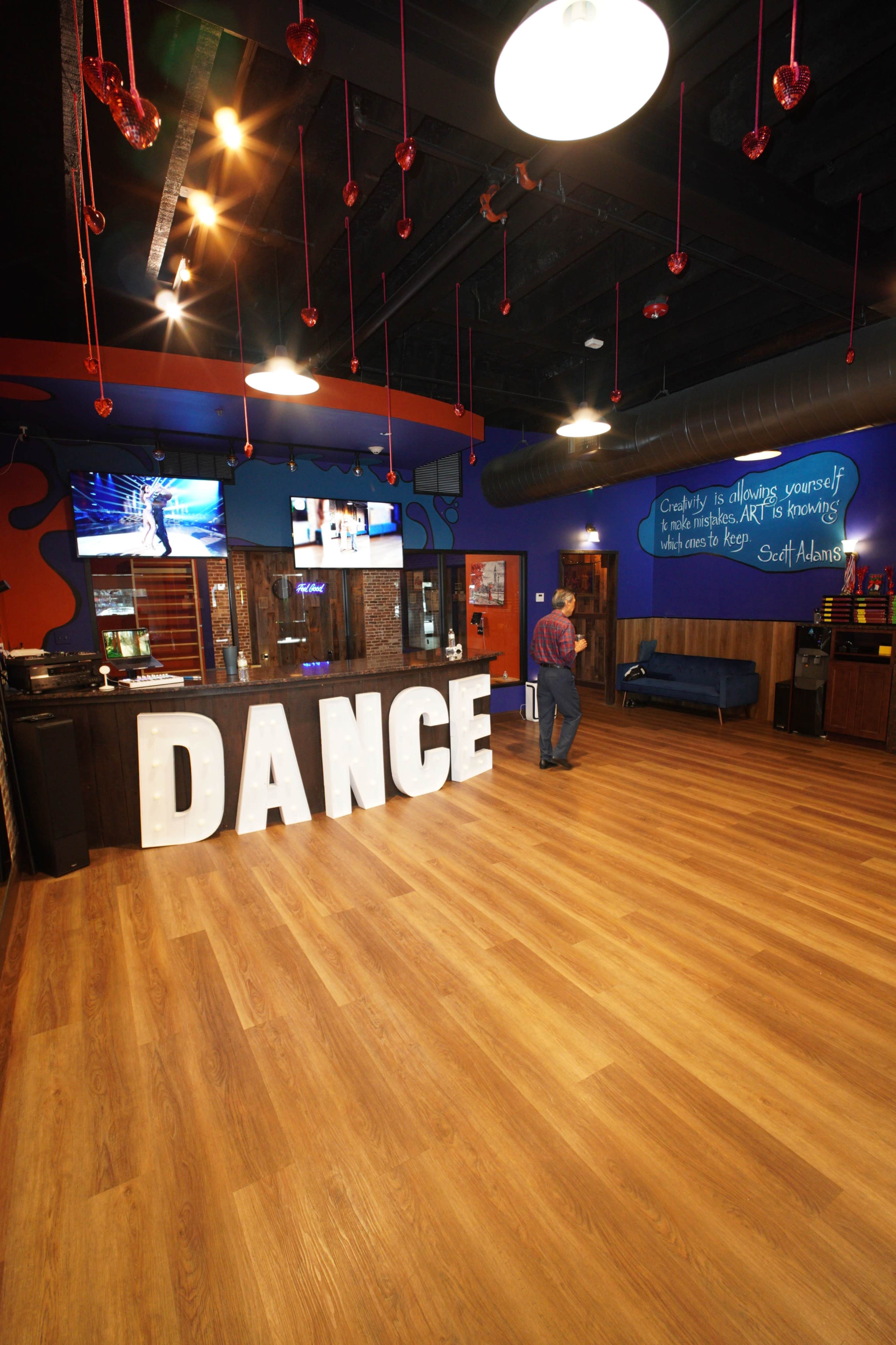 The image shows a spacious dance studio with large "DANCE" letters in front, a blue sofa in the background, and colorful walls adorned with artwork and a motivational quote.