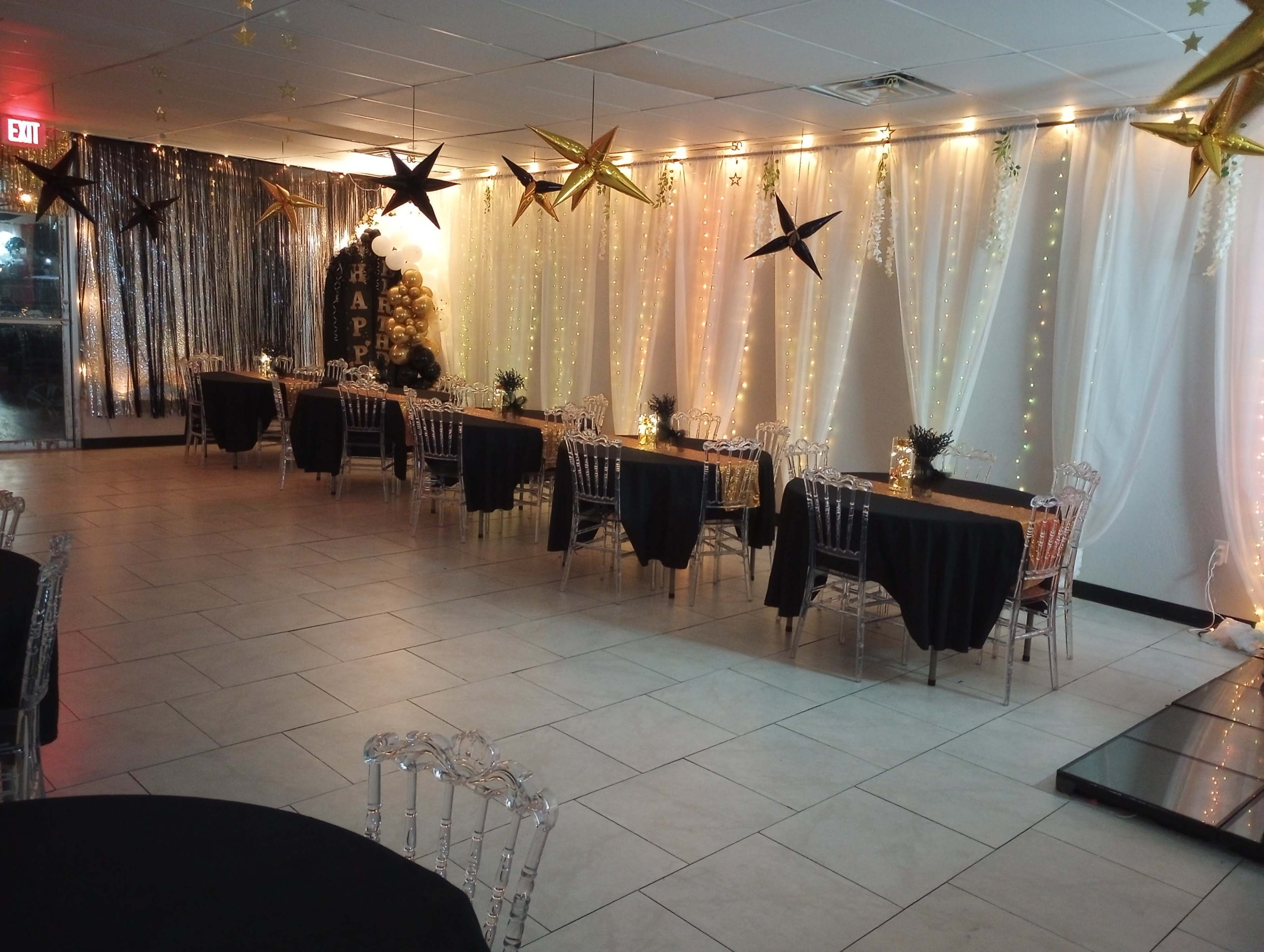The image shows a decorated event space with tables covered in black cloth, clear chairs, and golden stars hanging from the ceiling.