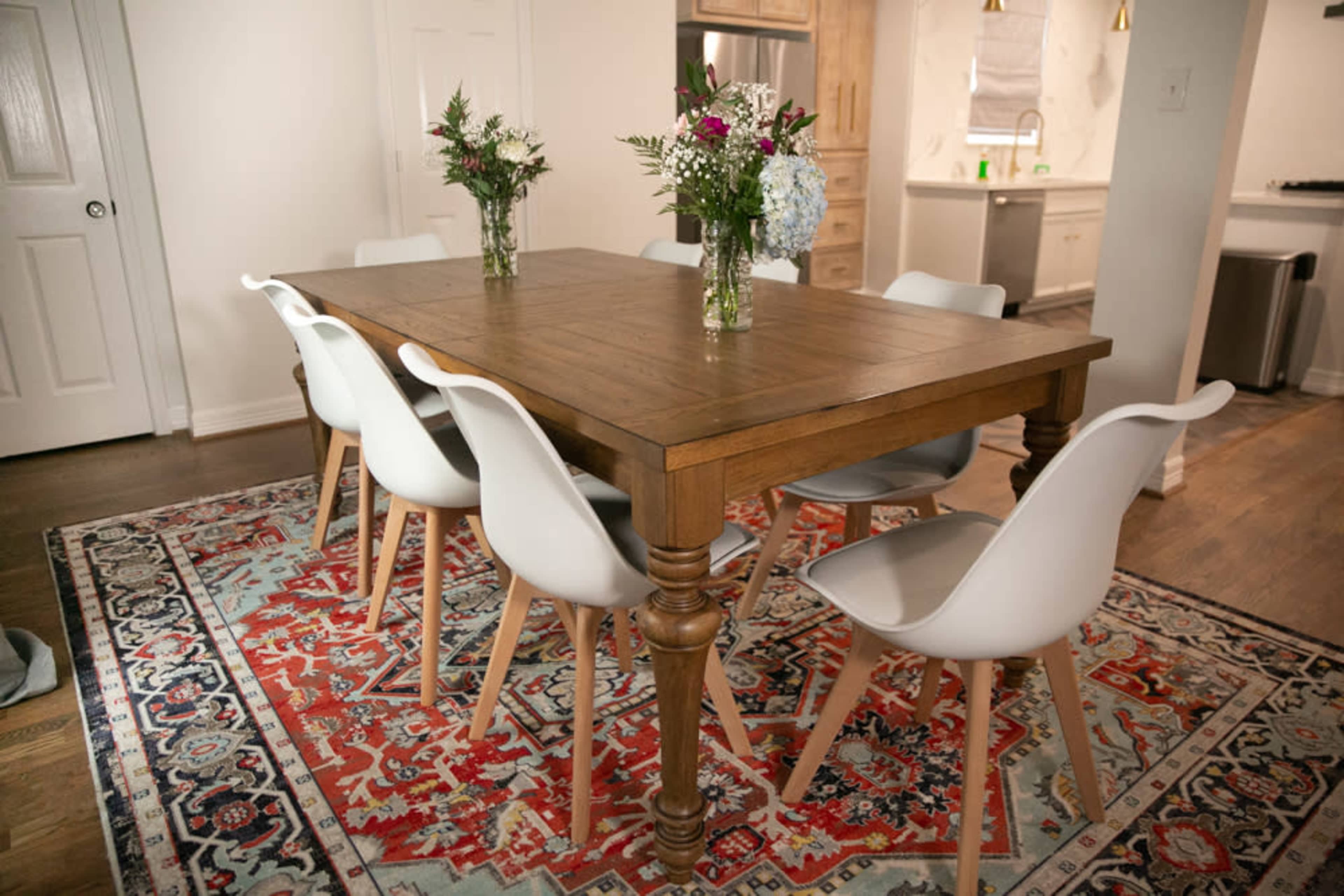A wooden dining table with a set of six white chairs is positioned on a patterned rug, flanked by vases of flowers.