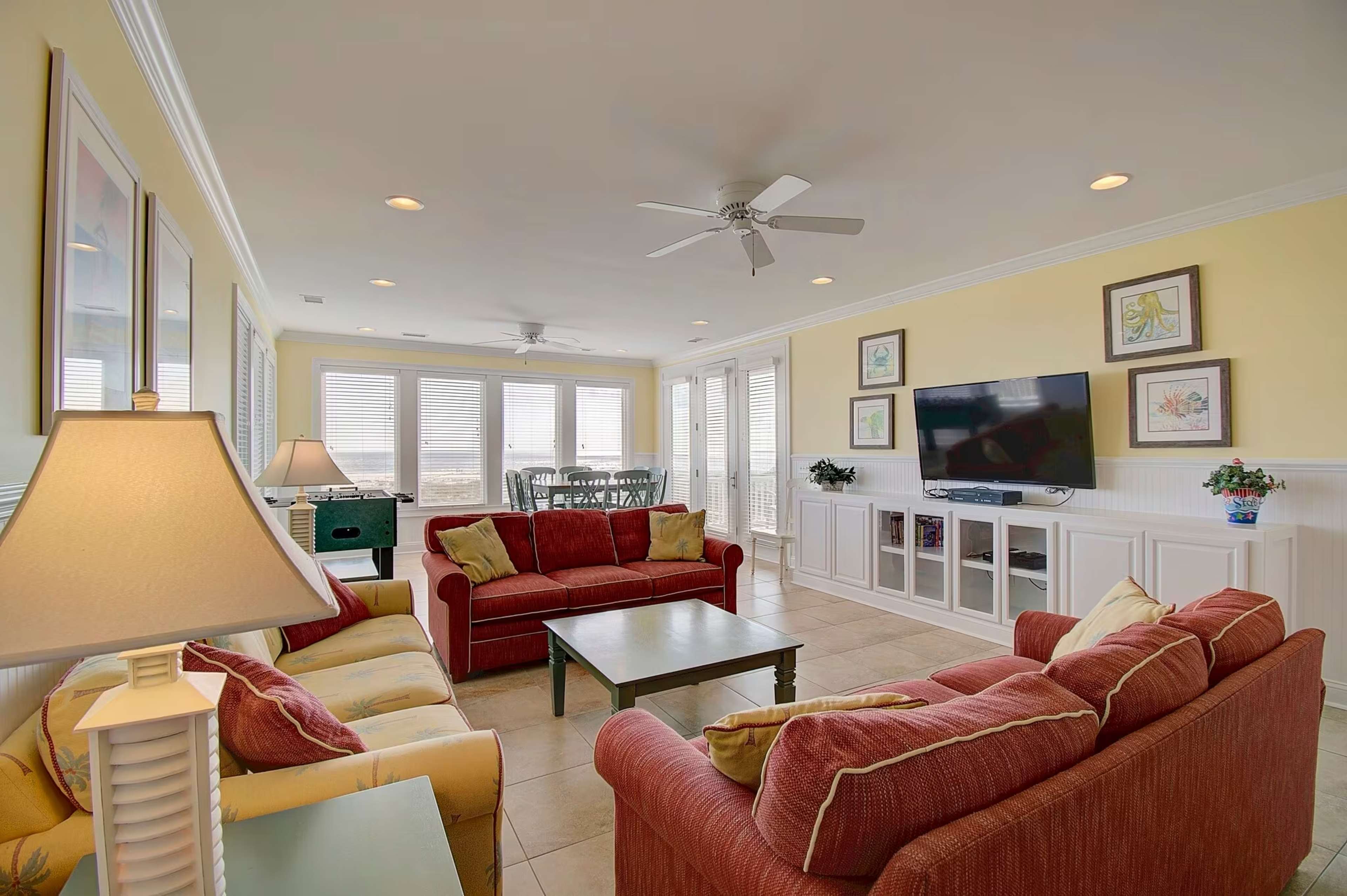 The living room features two red sofas, a green coffee table, a ceiling fan, and a wall-mounted television, with large windows providing natural light.
