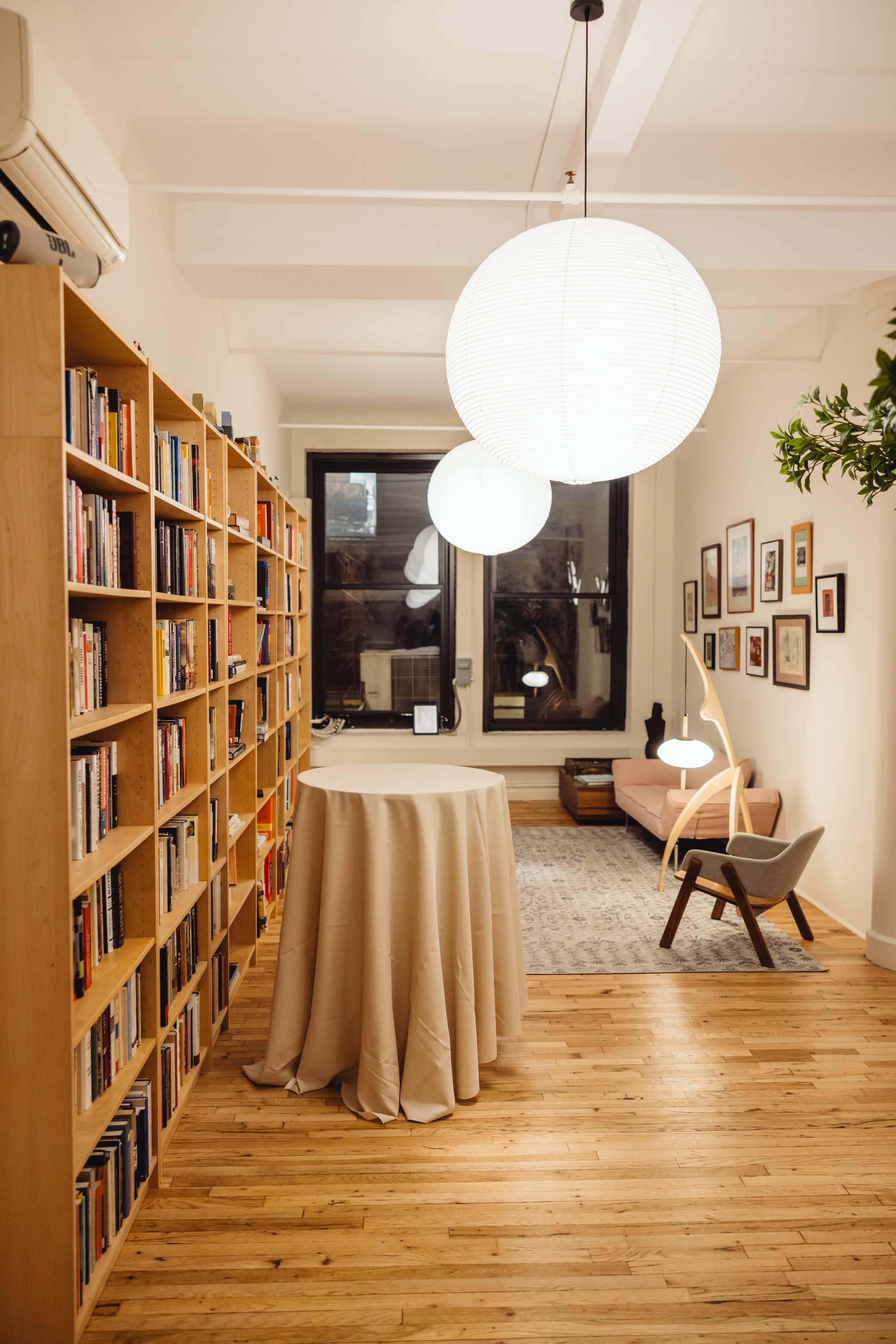 The image shows a cozy room featuring a large bookshelf filled with books, a table with a tablecloth, a pink sofa, and several framed pictures on the walls, illuminated by hanging light fixtures.