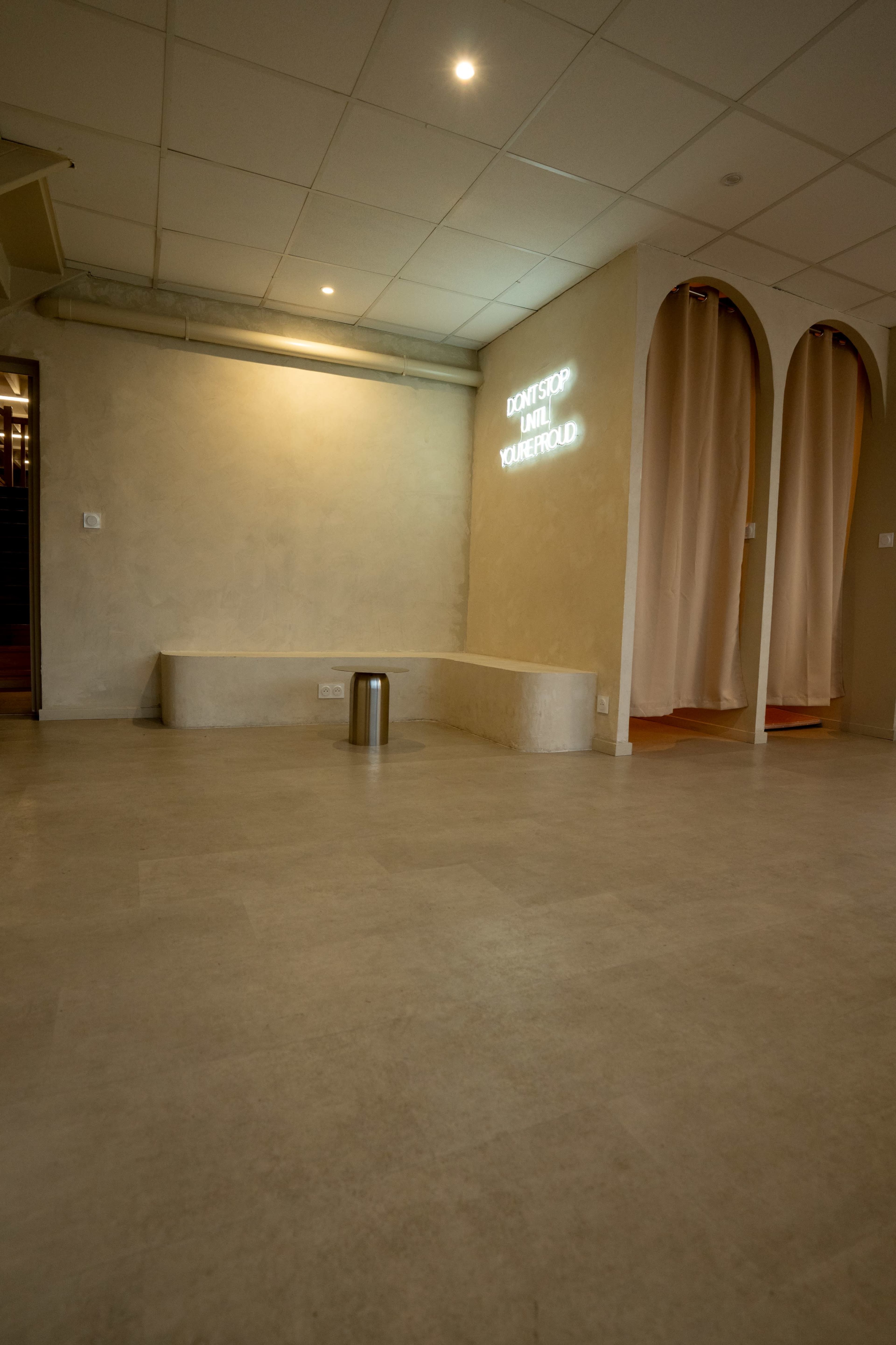 The image shows an empty interior space with a light-colored wall, a curved seating area, and a metal trash can, illuminated by a neon sign that reads "DON'T STOP UNTIL YOU'RE PROUD."