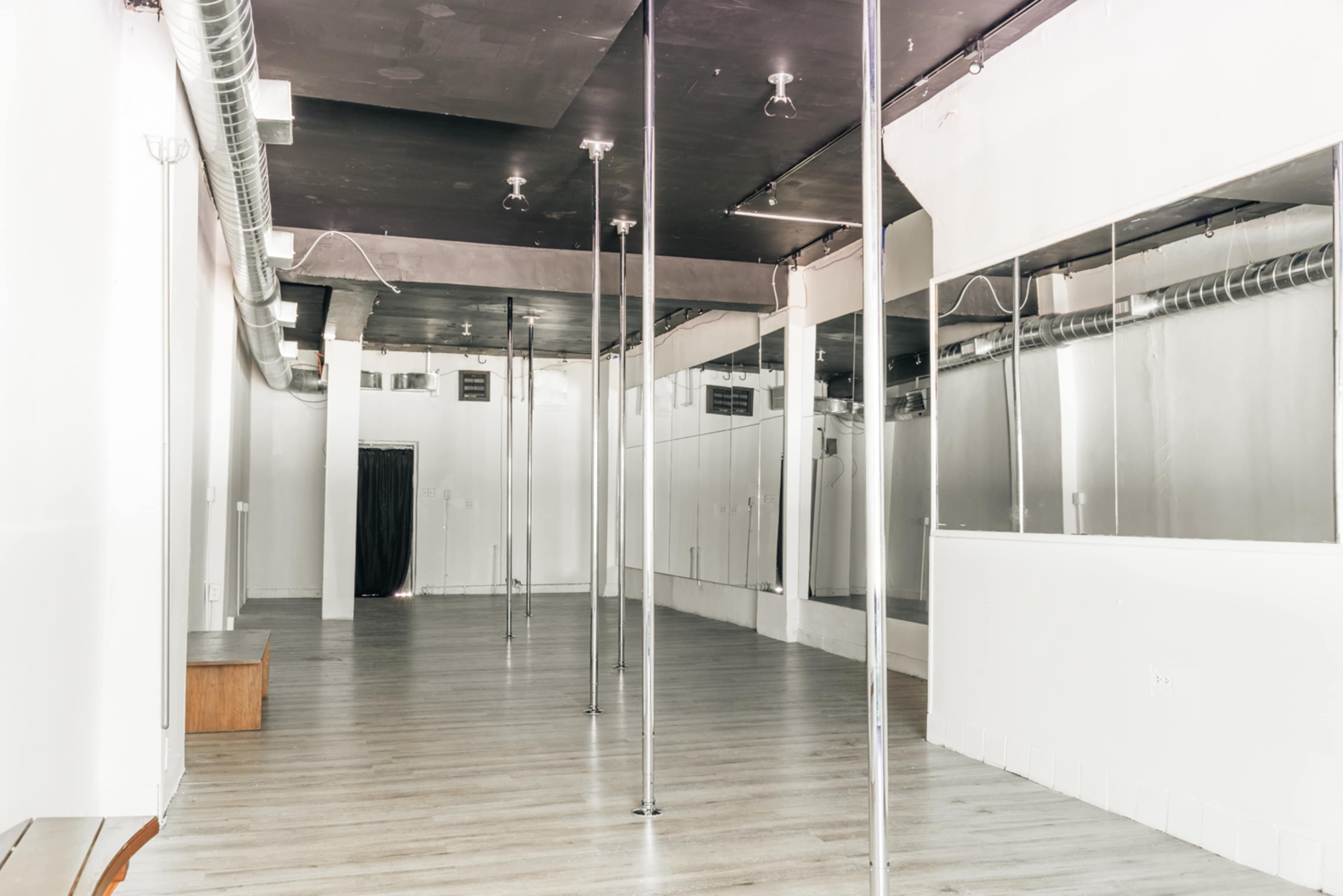 An empty dance studio with polished wooden floors, mirrored walls, and several vertical poles.