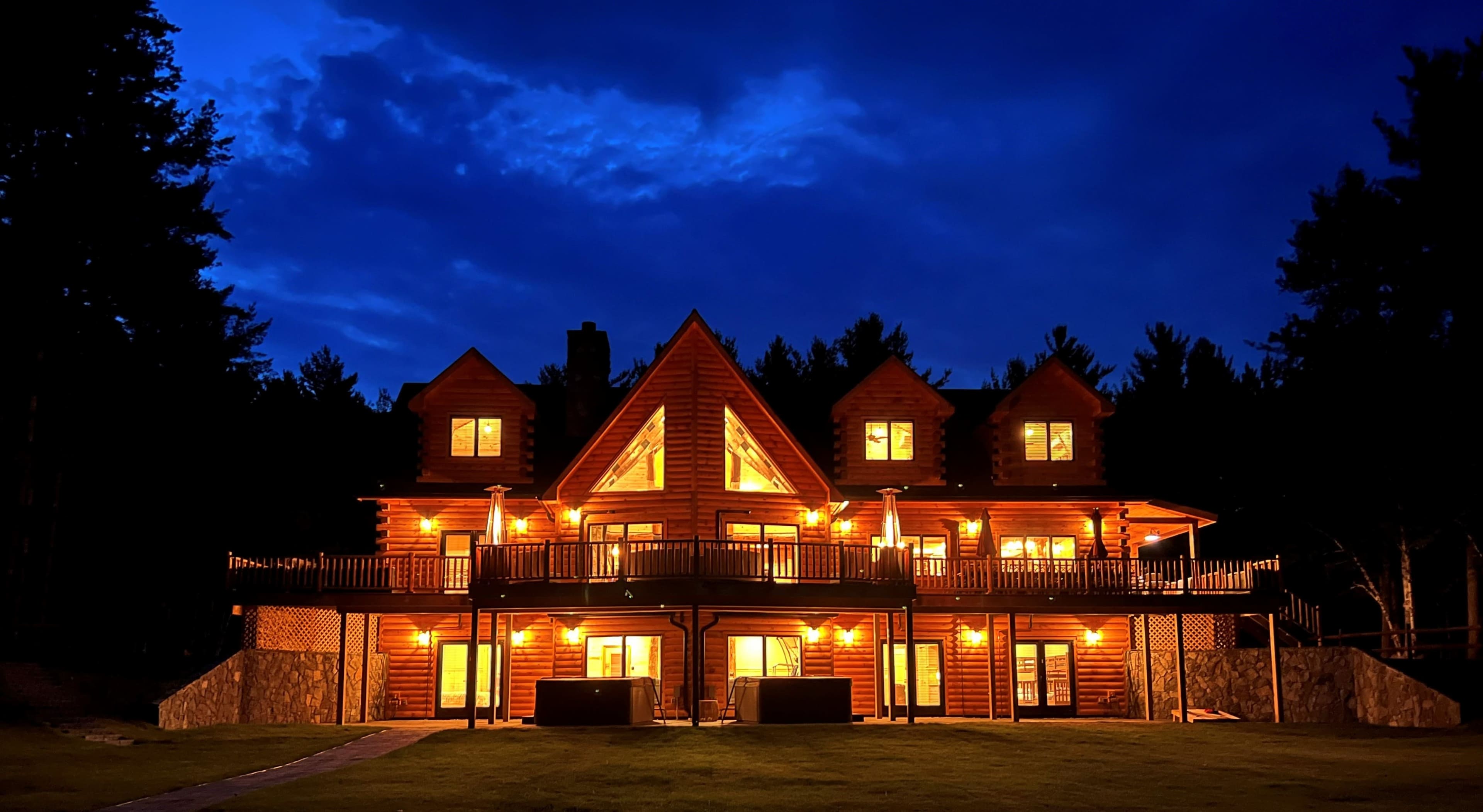A spacious log cabin with multiple gabled roofs is illuminated by outdoor lights against a twilight sky.