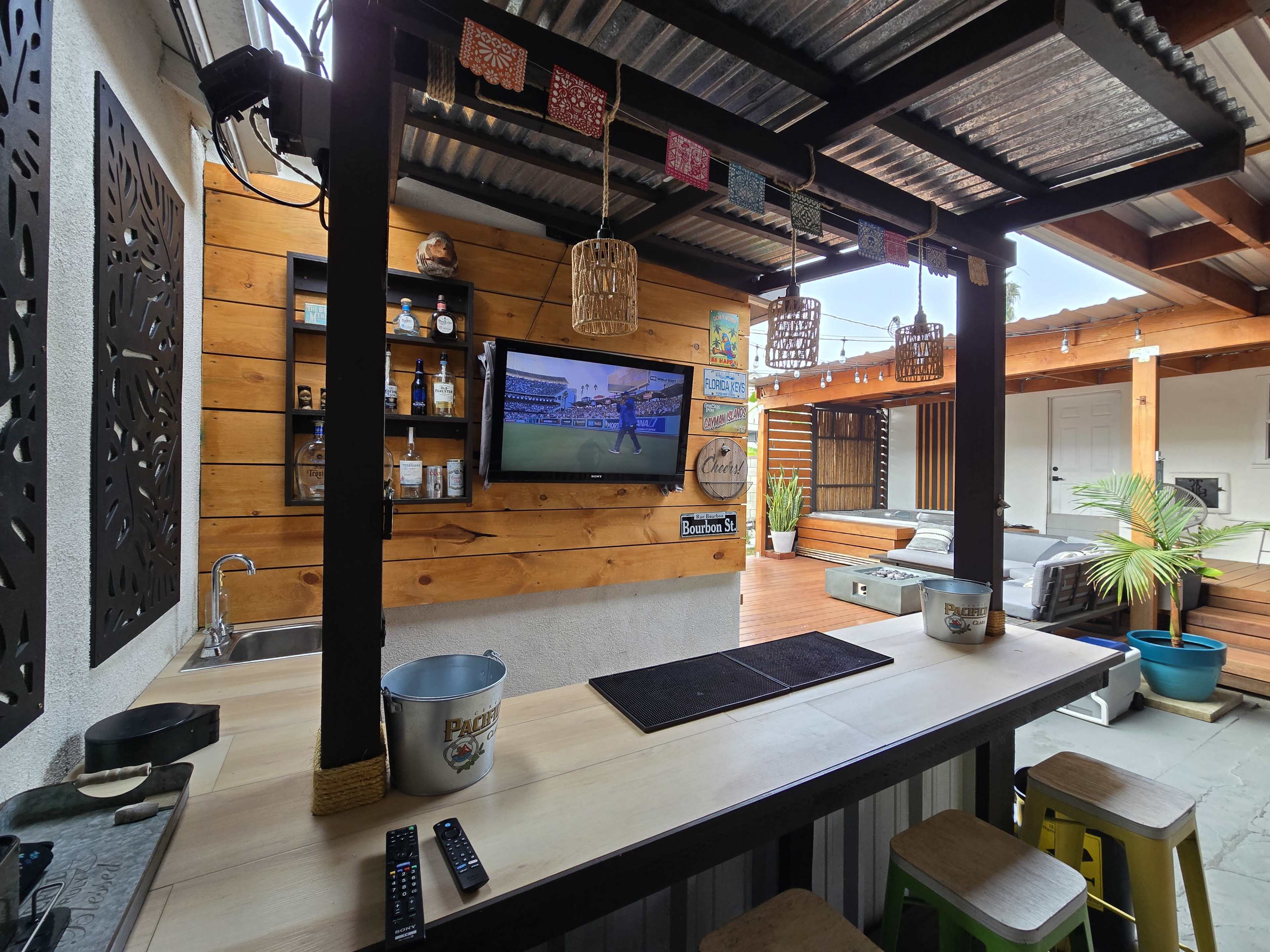 The image shows a rustic outdoor bar area with a wooden countertop, a television mounted on the wall, shelves displaying bottles, and colorful decorations hanging above.