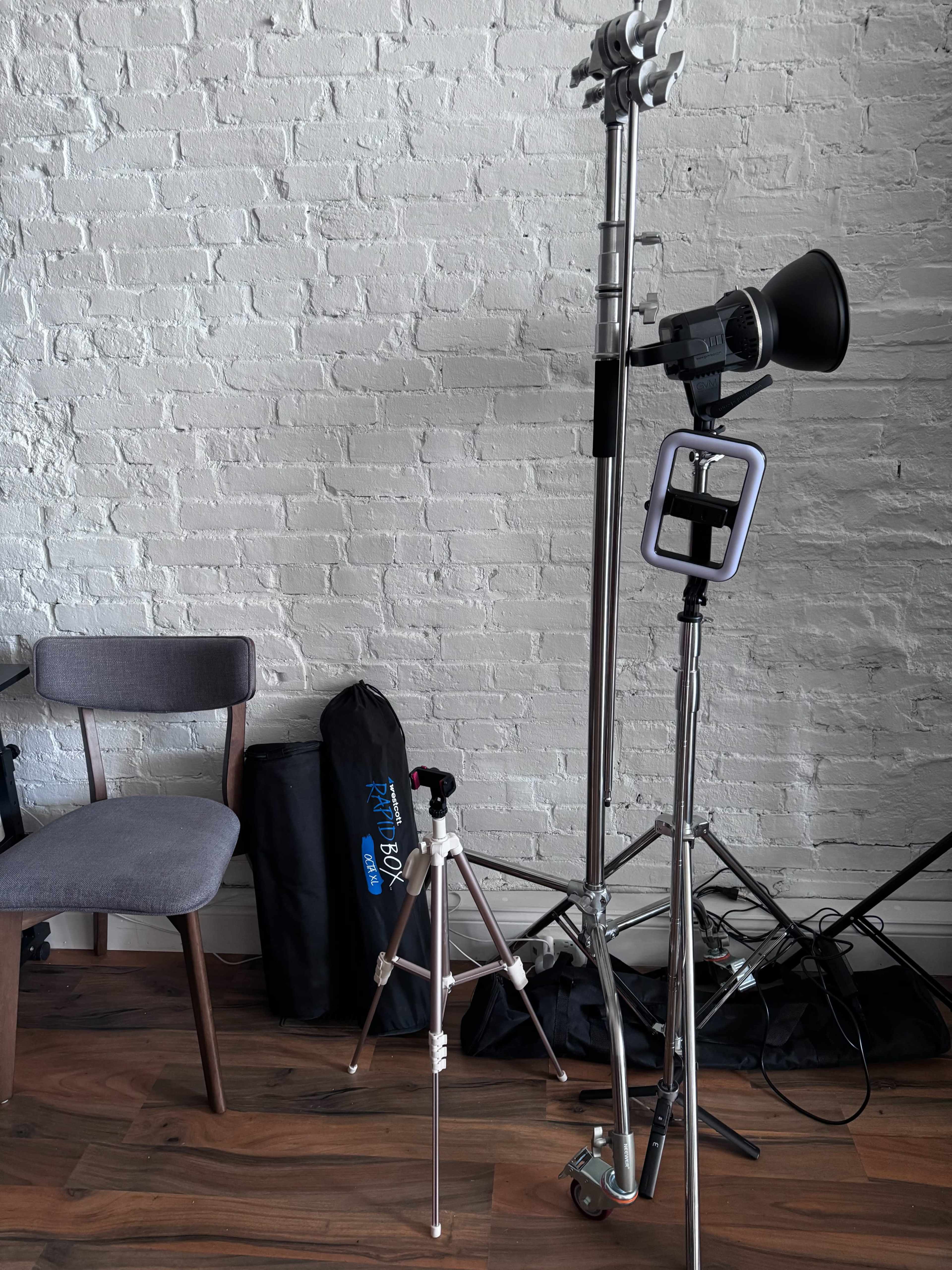 The image shows a photography setup with lighting equipment, a tripod, and a gray chair in front of a brick wall.