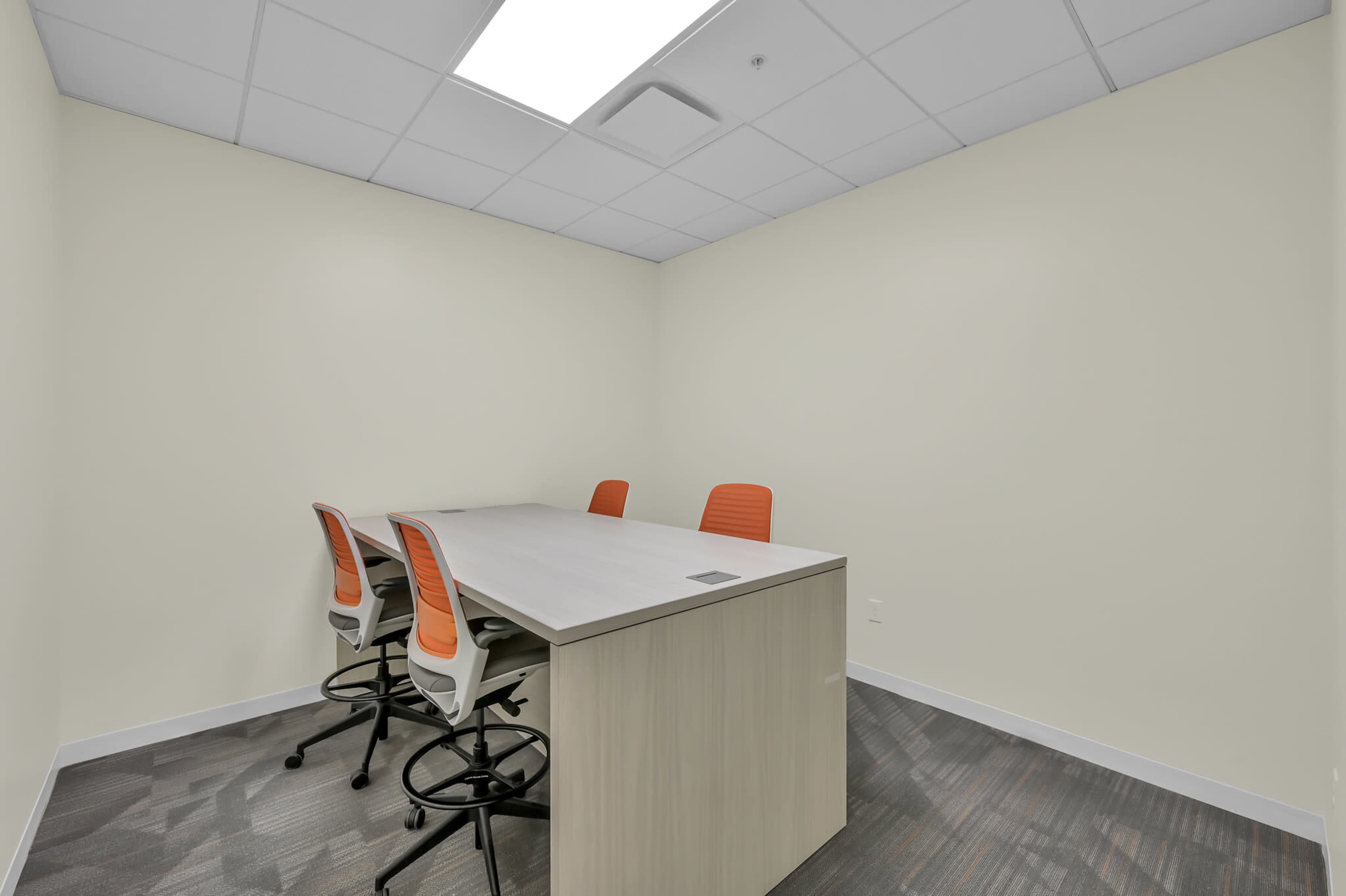 A meeting room features a rectangular table surrounded by four high-back chairs with orange seats.