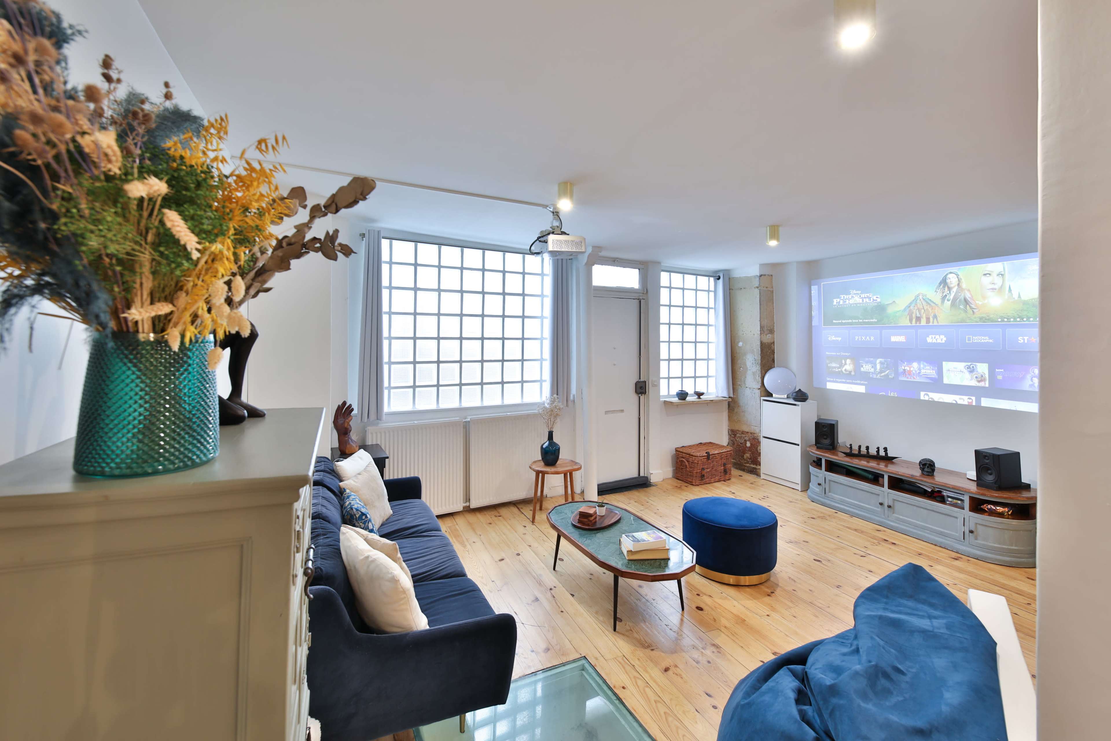 A bright, modern living room with a projector screen displaying content on one wall, a cozy sofa, and large windows letting in natural light.