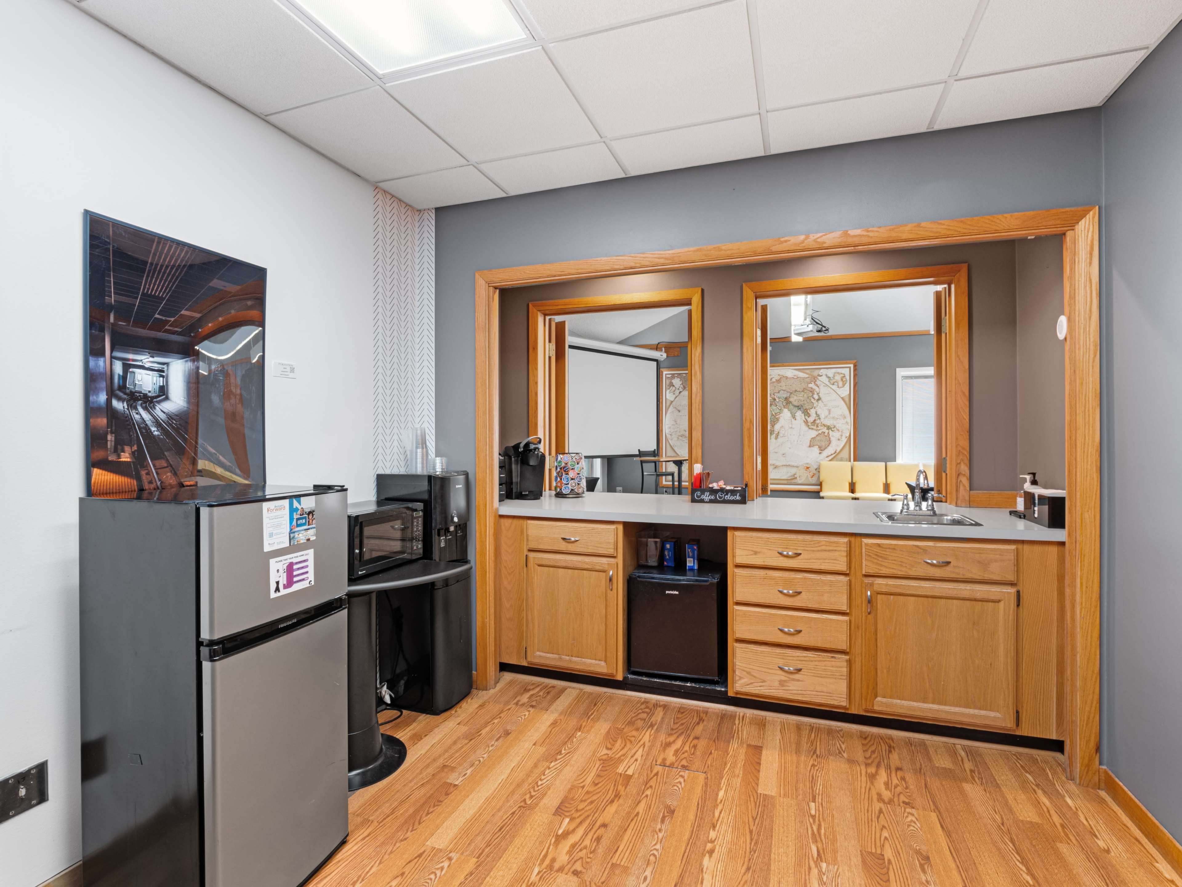 A kitchenette area with wooden cabinets, a small refrigerator, a microwave, and a countertop, separated from a room with a map on the wall.