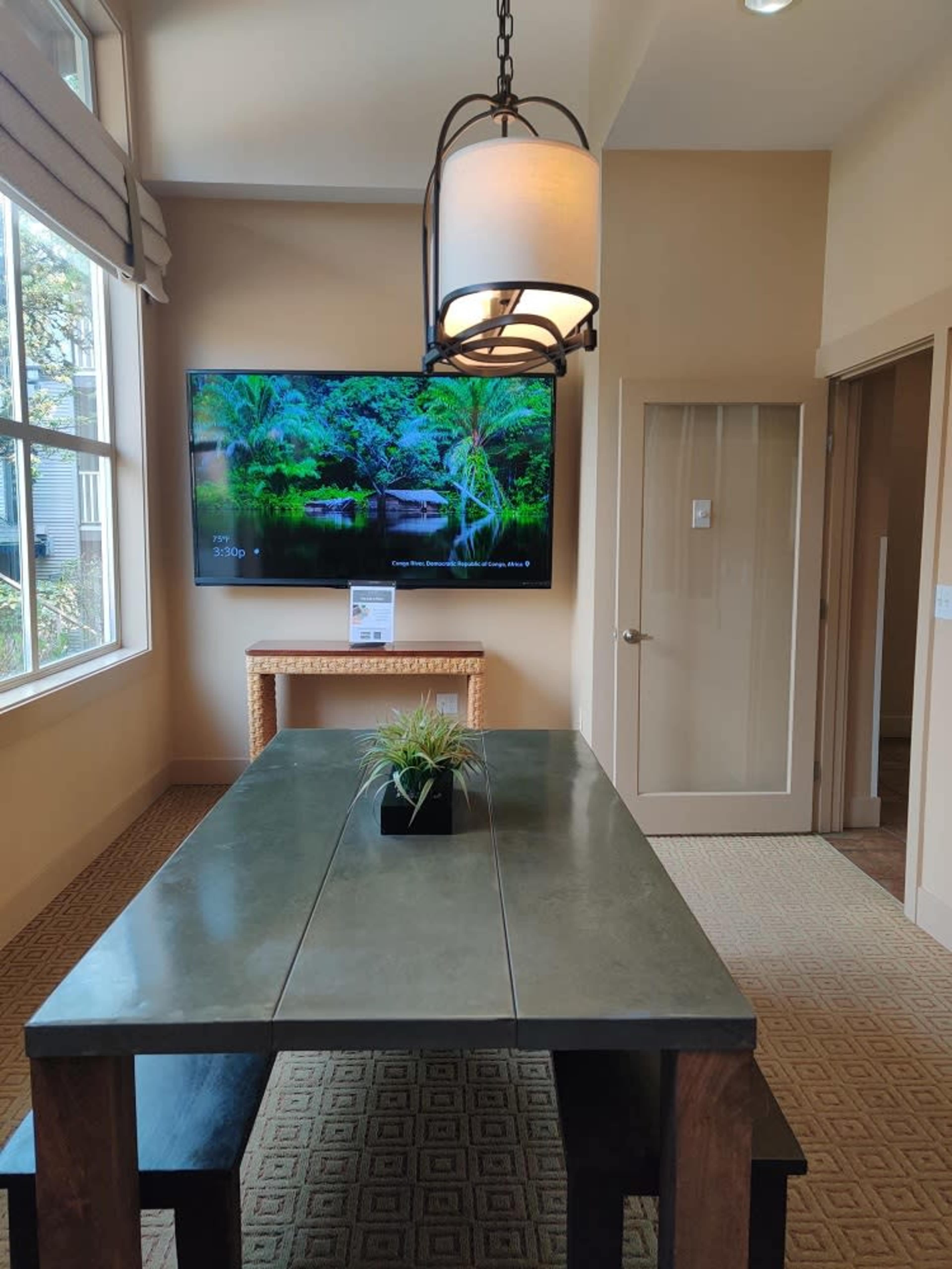 A rectangular table is positioned in a room with large windows and a wall-mounted television displaying a nature scene.