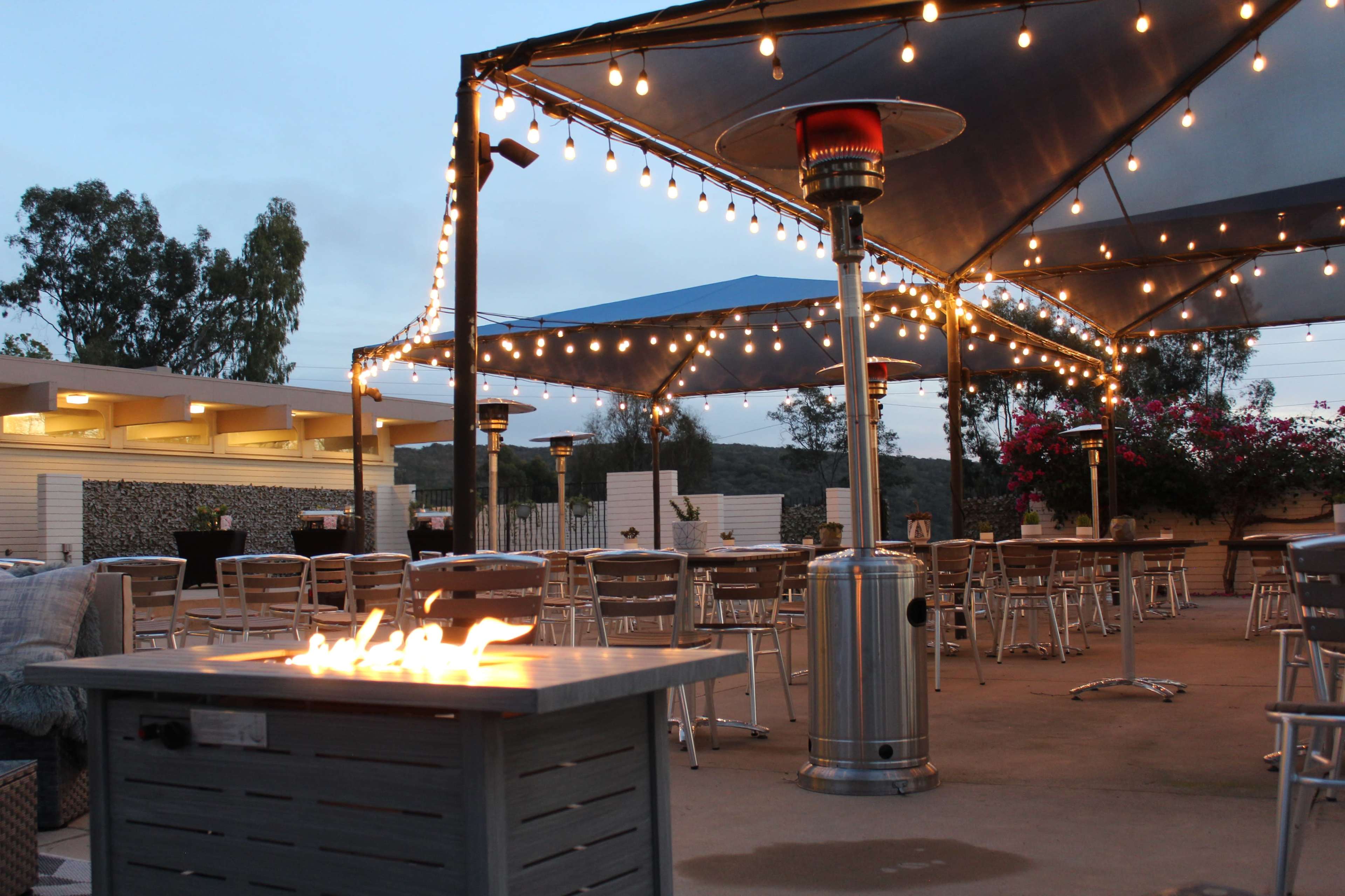 An outdoor dining area features several tables under lit canopies, with a fire pit in the foreground.