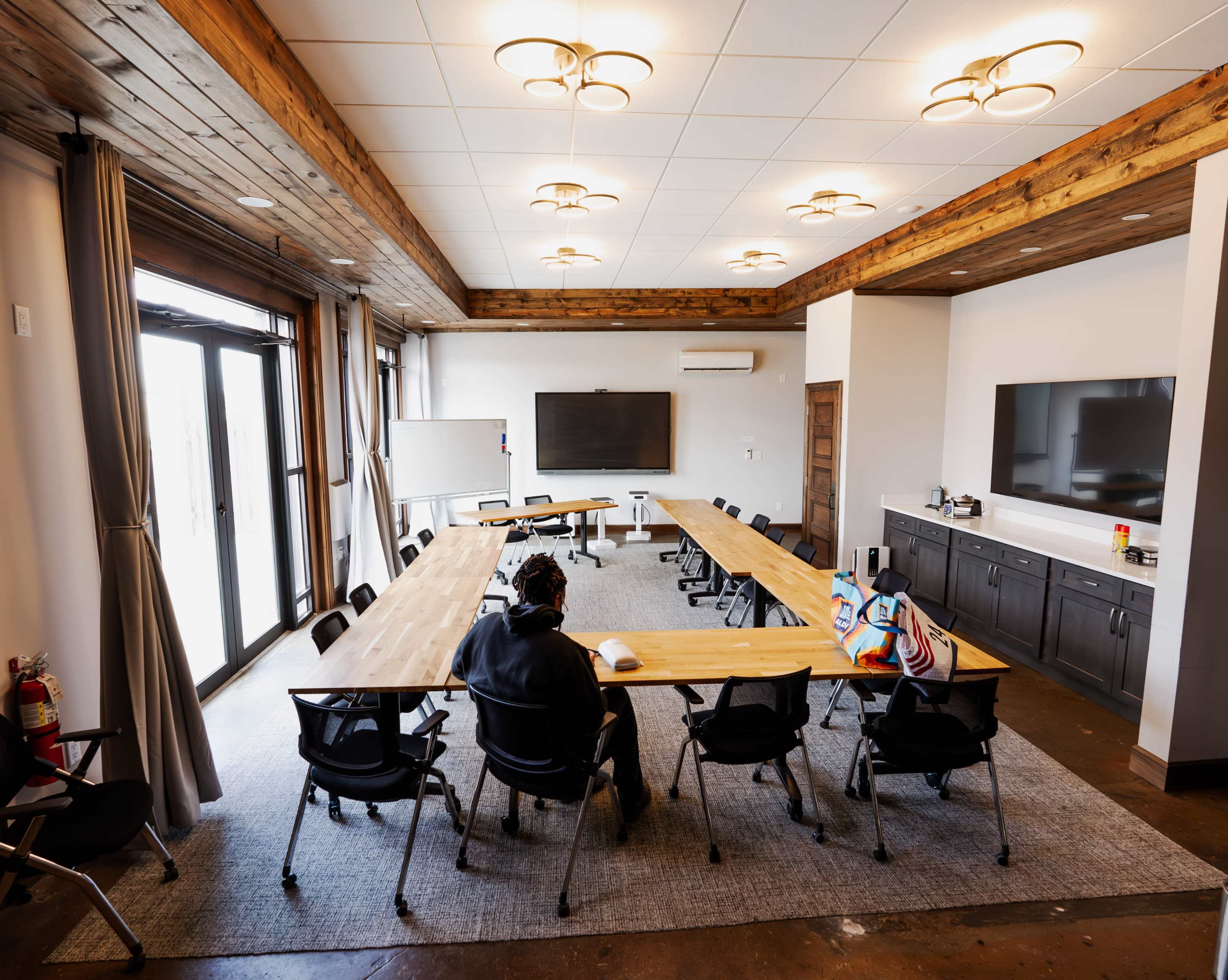 A modern conference room features a long wooden table surrounded by black chairs, with large windows and two screens mounted on the walls.