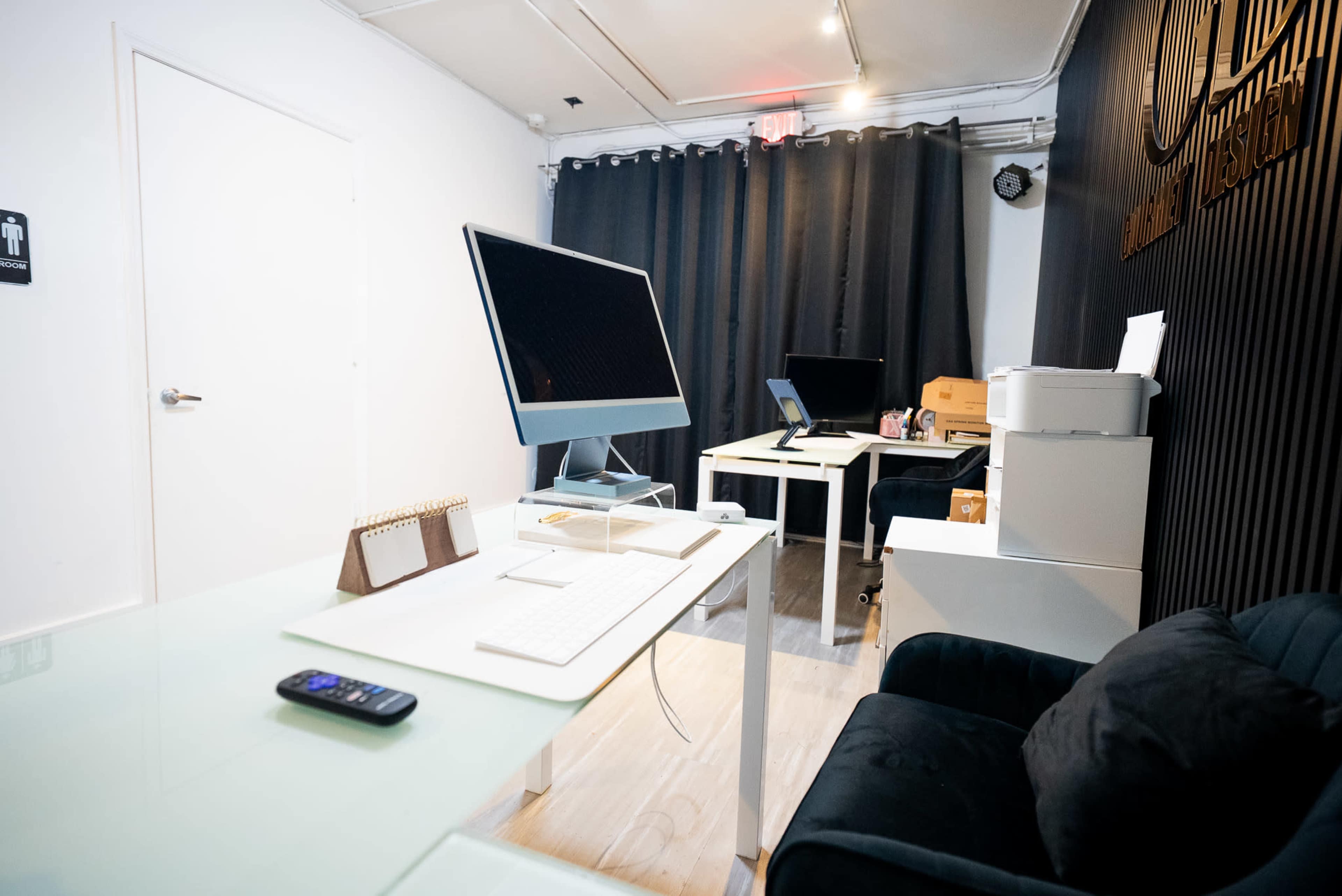 The image shows a modern office space with two desks, one equipped with an Apple computer, and a seating area featuring a black couch.