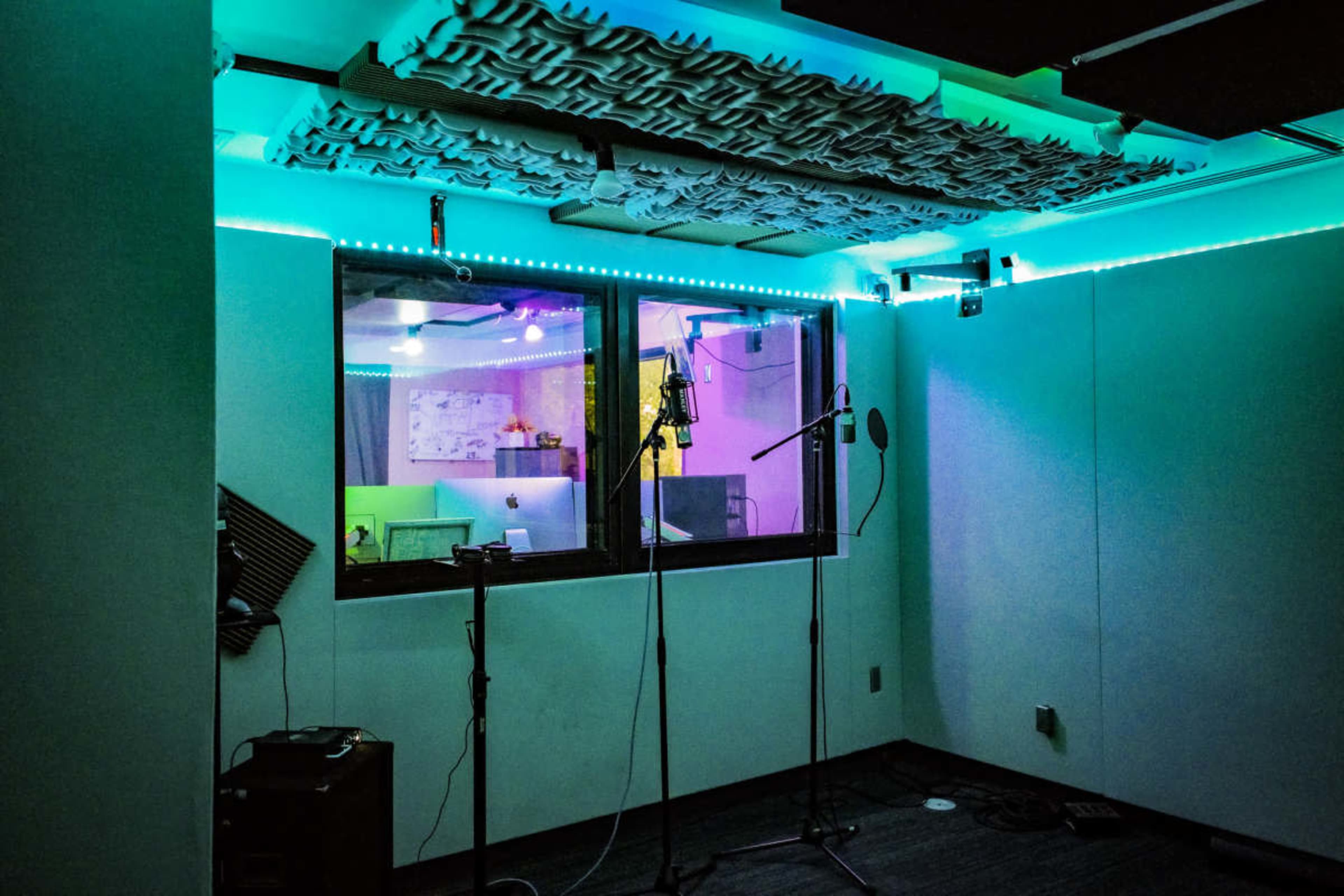 The image shows a sound studio with microphones set up in front of a glass window, illuminated by colored LED lights.