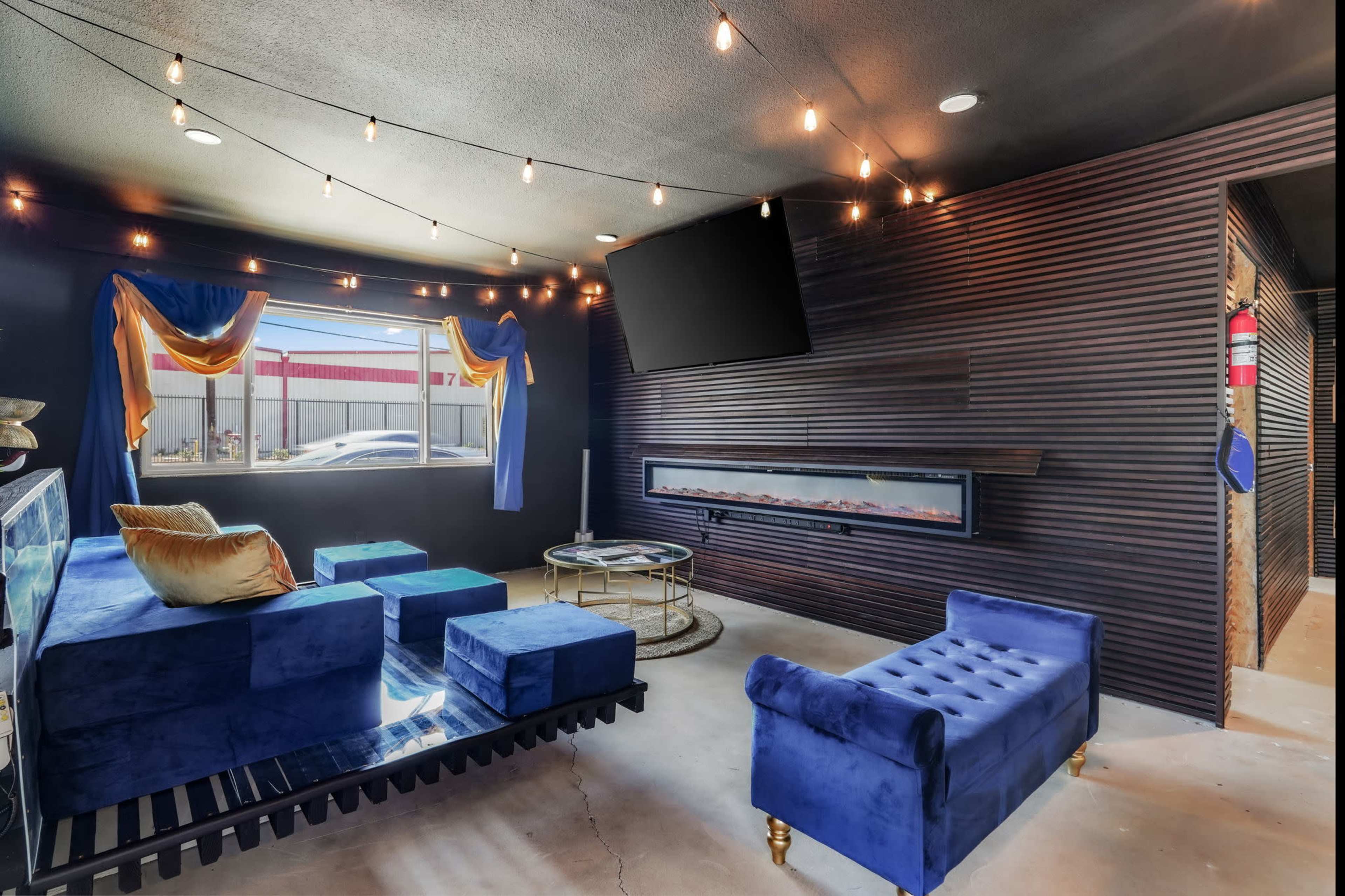 The image shows a modern lounge area featuring blue velvet furniture, a wall-mounted TV, and an electric fireplace against a dark paneled wall.