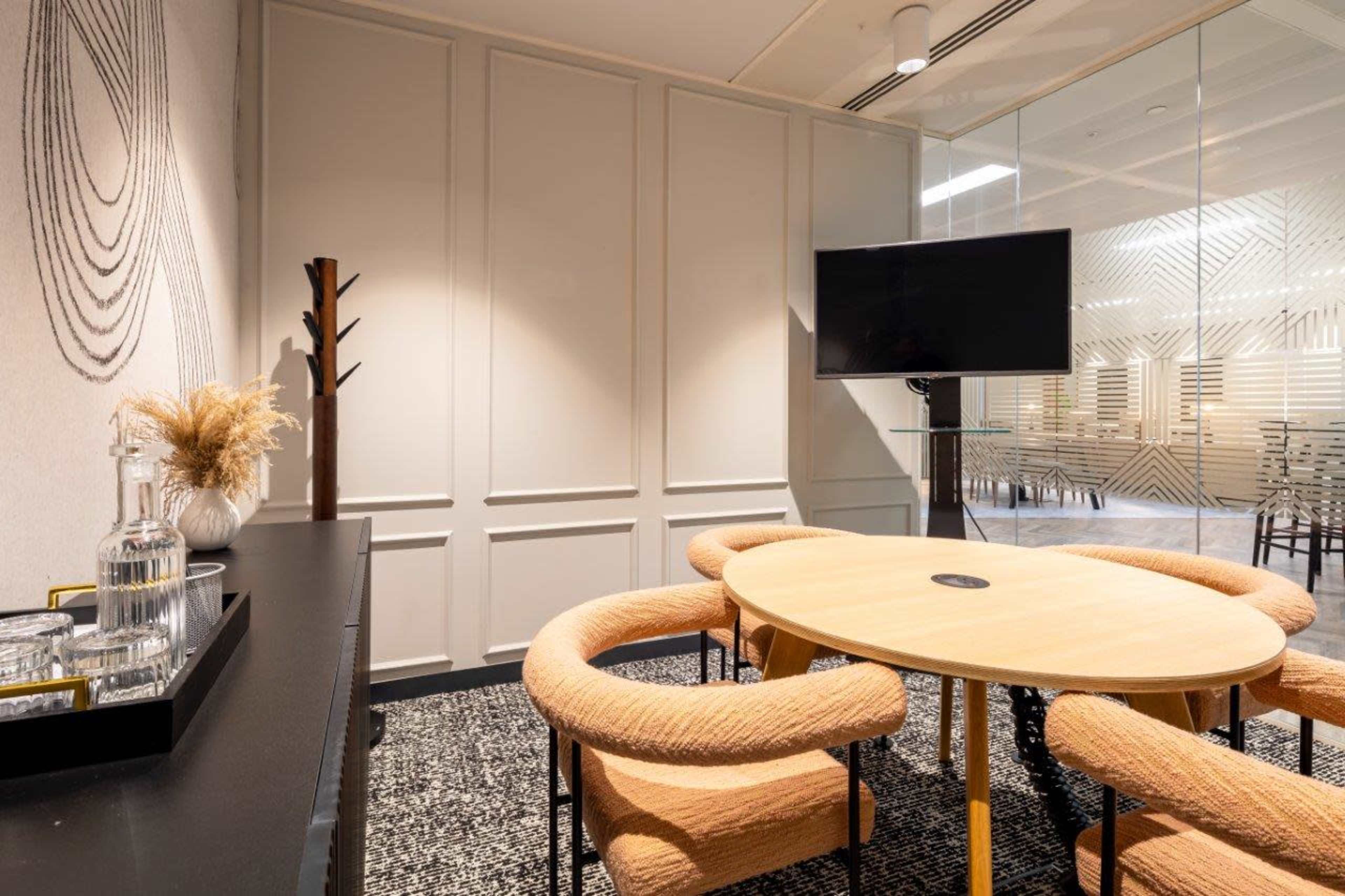 A modern conference room with a round wooden table surrounded by upholstered chairs, a glass wall in the background, and a television mounted on the wall.