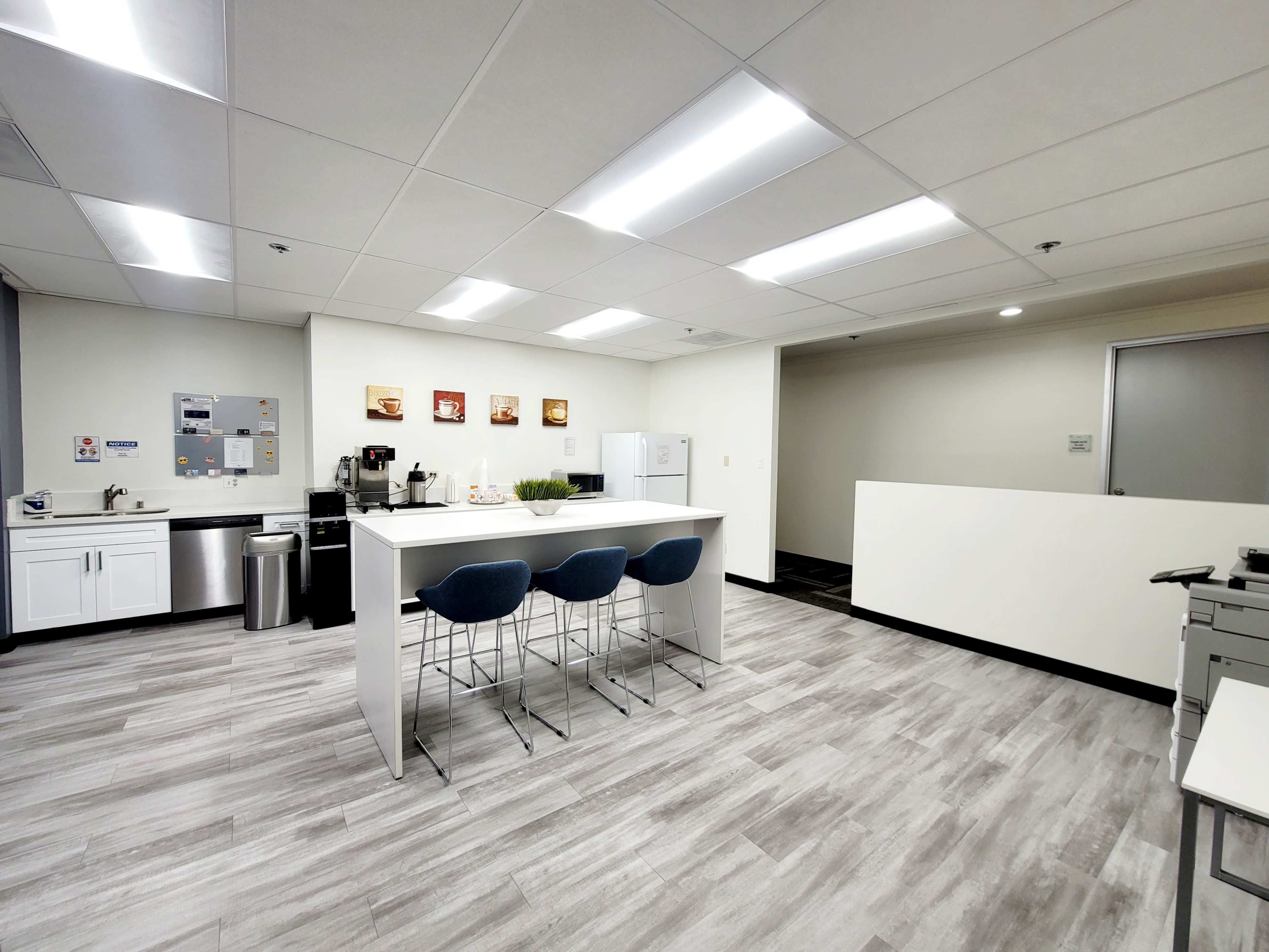 A modern break room with a kitchen area featuring a counter, stools, coffee machine, and appliances, set in an open office environment.