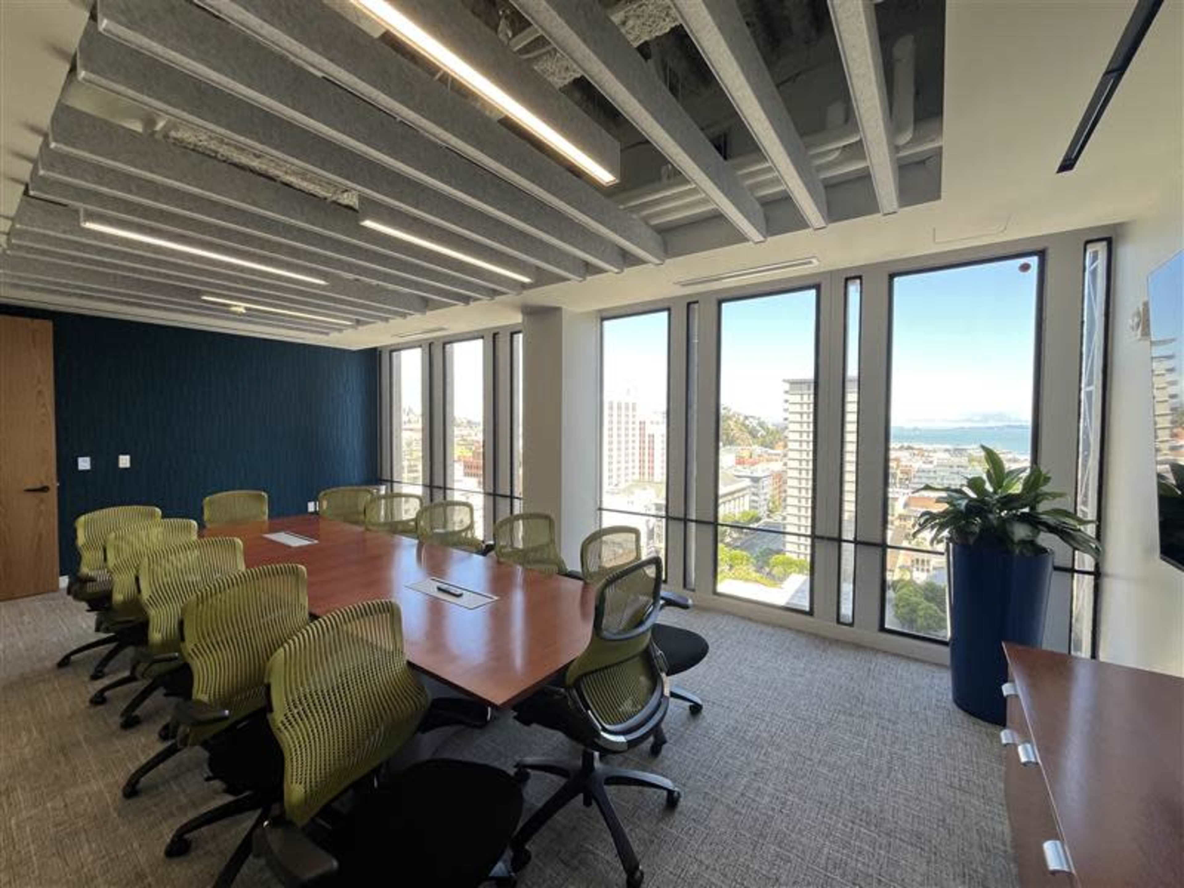 A modern conference room features a large wooden table surrounded by green office chairs, with floor-to-ceiling windows offering a view of the city.