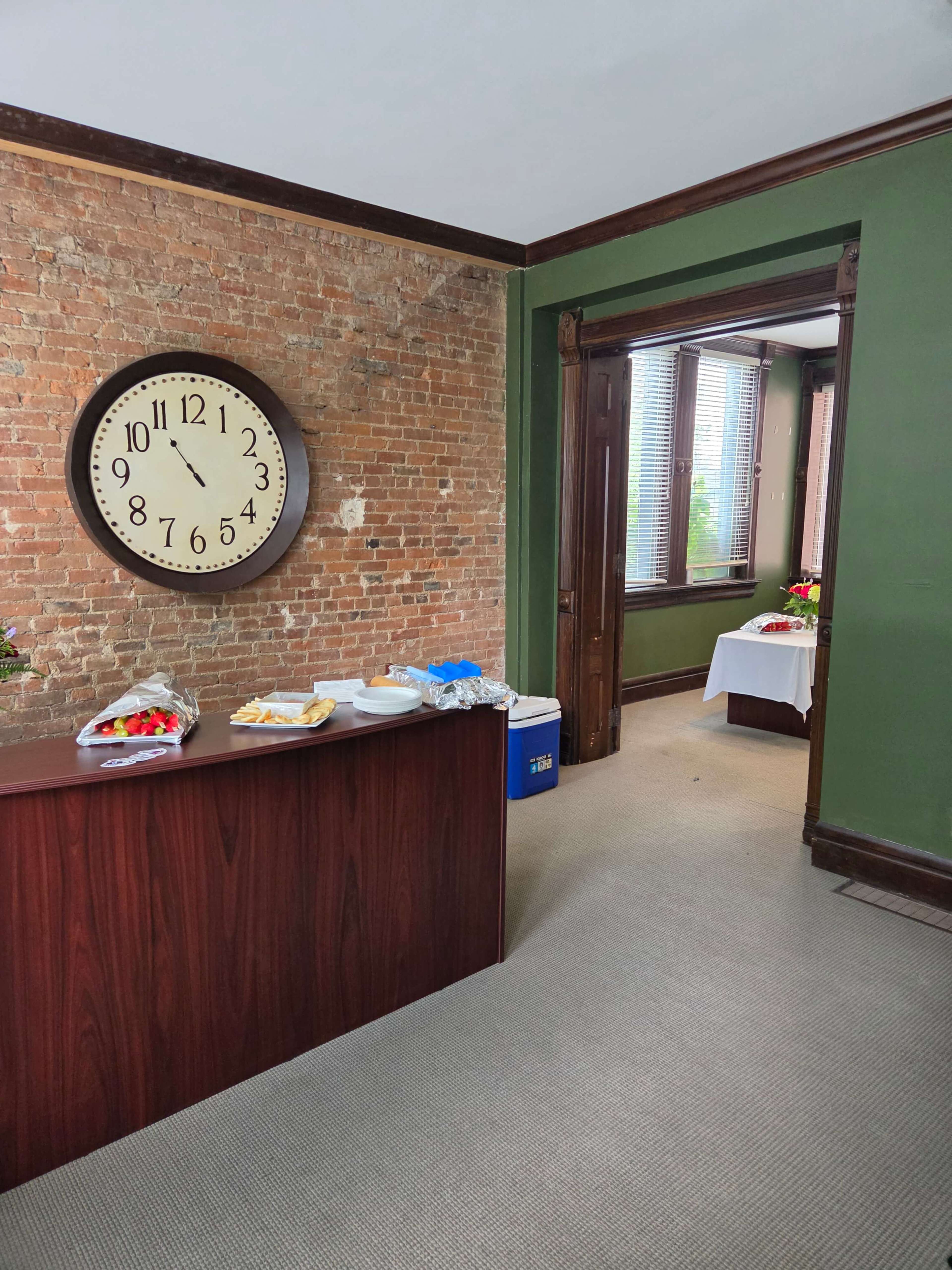 A wooden reception desk stands against a brick wall, with a large clock on display, while an open doorway leads to another room featuring tables covered with white tablecloths.