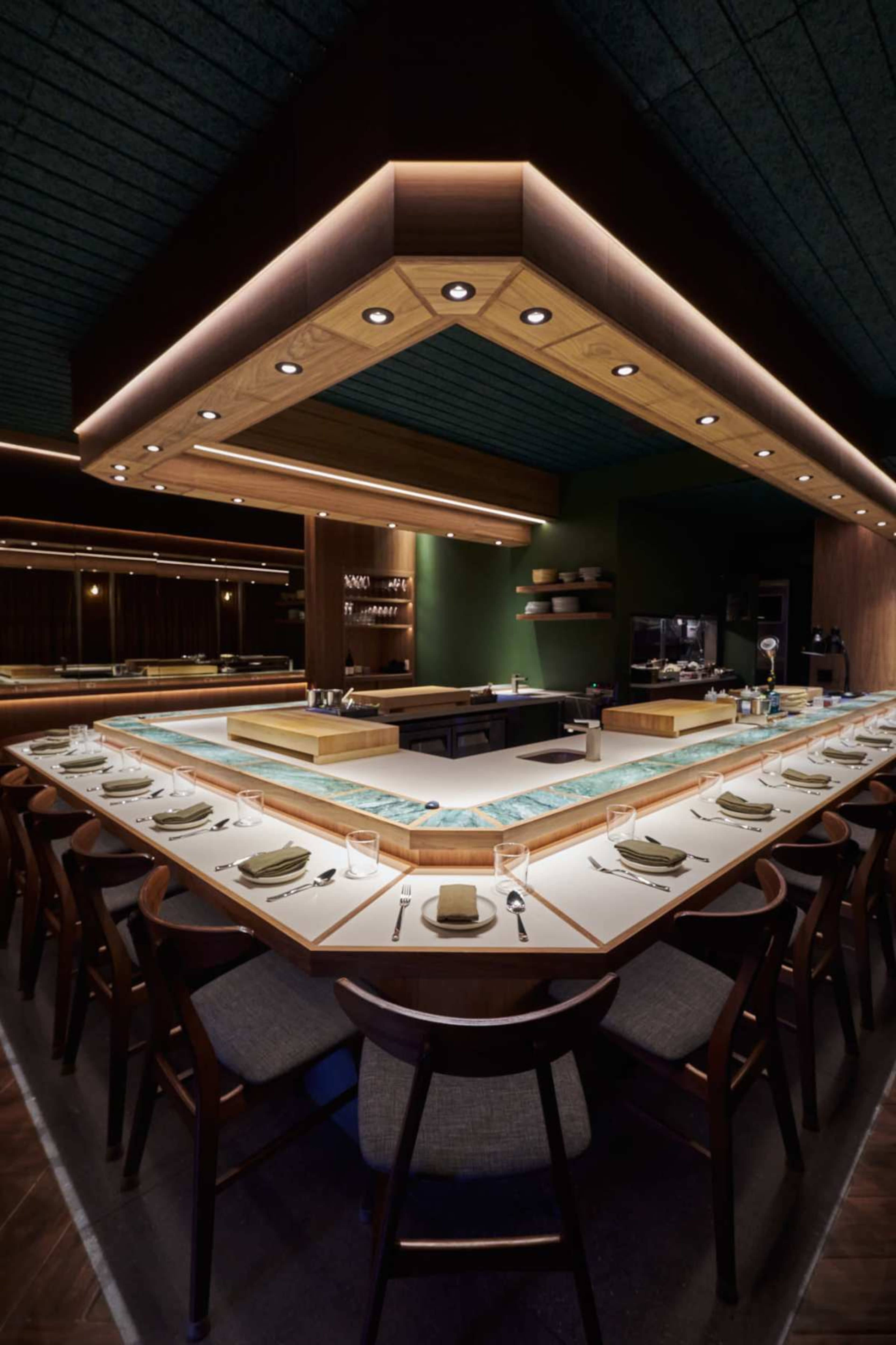 The image shows a modern, open-concept restaurant interior featuring a central dining counter with seating around it and warm lighting illuminating the space.
