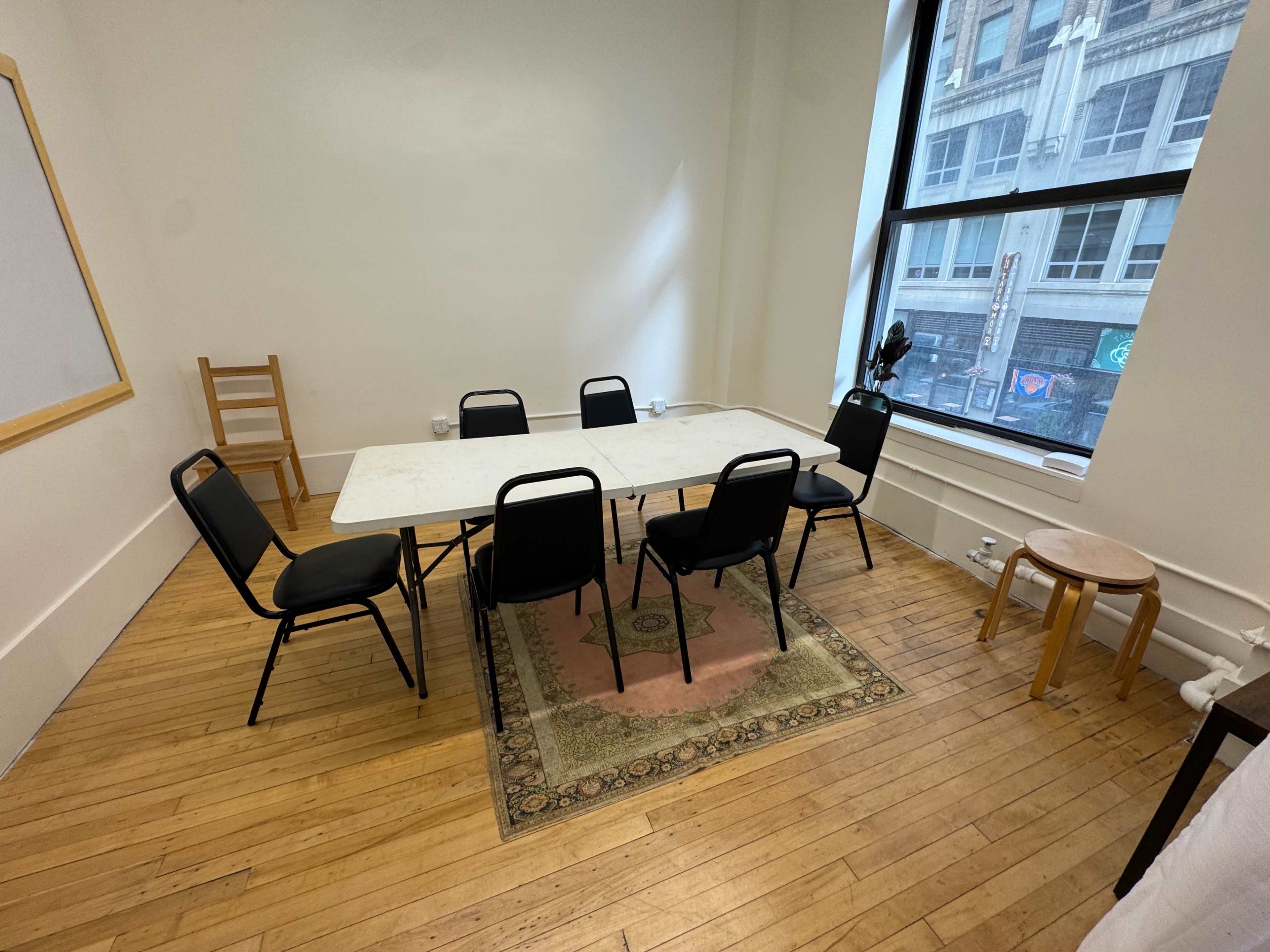The image shows a small, sparse meeting room with a large table surrounded by six black chairs, a rug under the table, and a wooden stool in the corner.