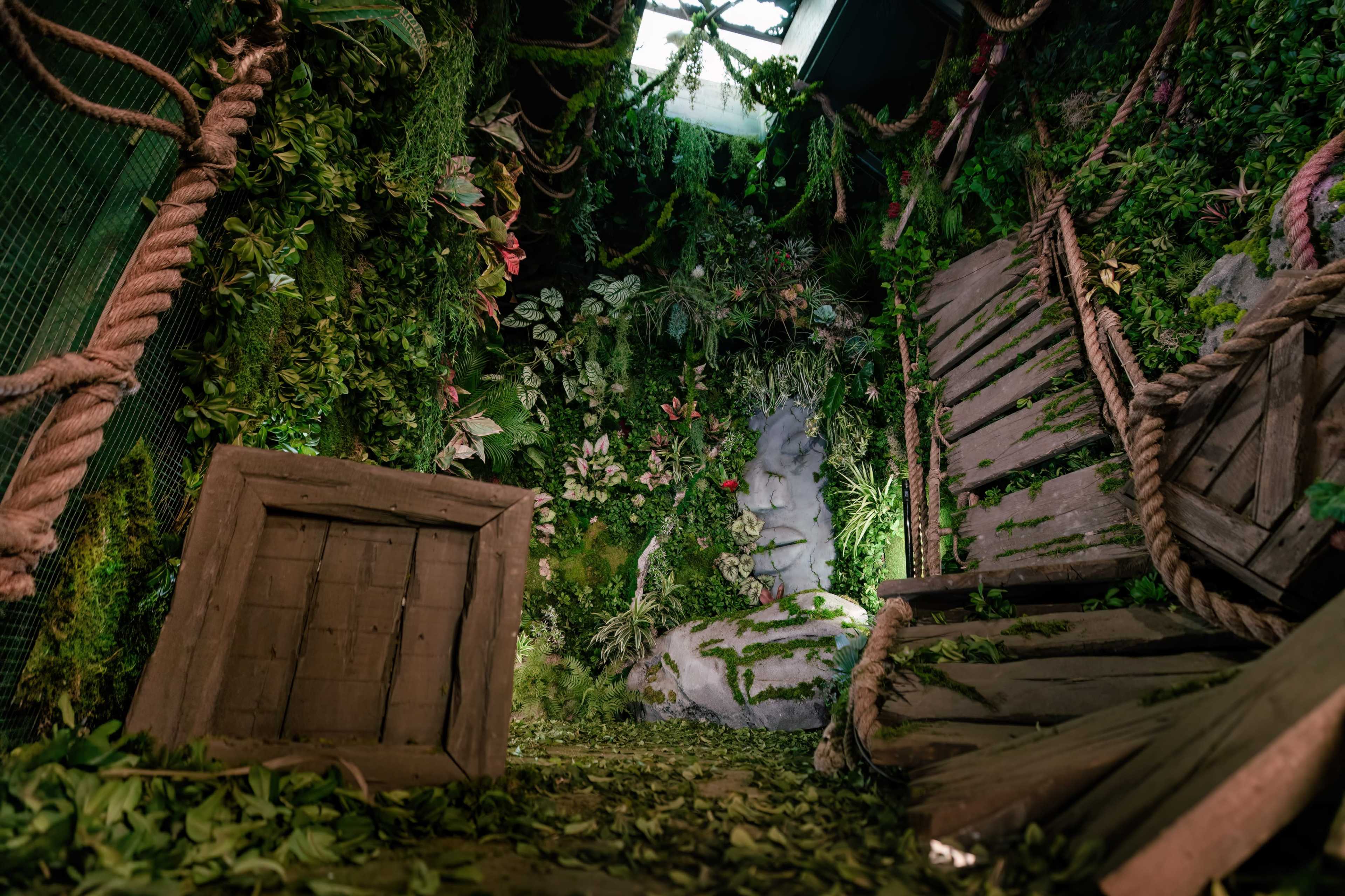 A room filled with lush greenery, featuring a stone figure and wooden structures that appear to be in disarray.