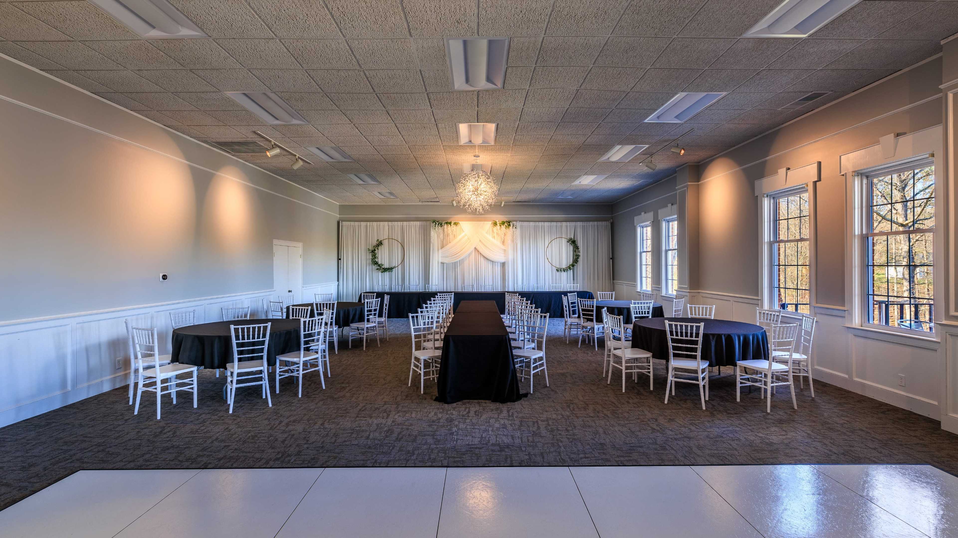 A spacious event room features several round tables with black tablecloths and white chiavari chairs arranged around a central black table, with decorative drapes and a chandelier above.