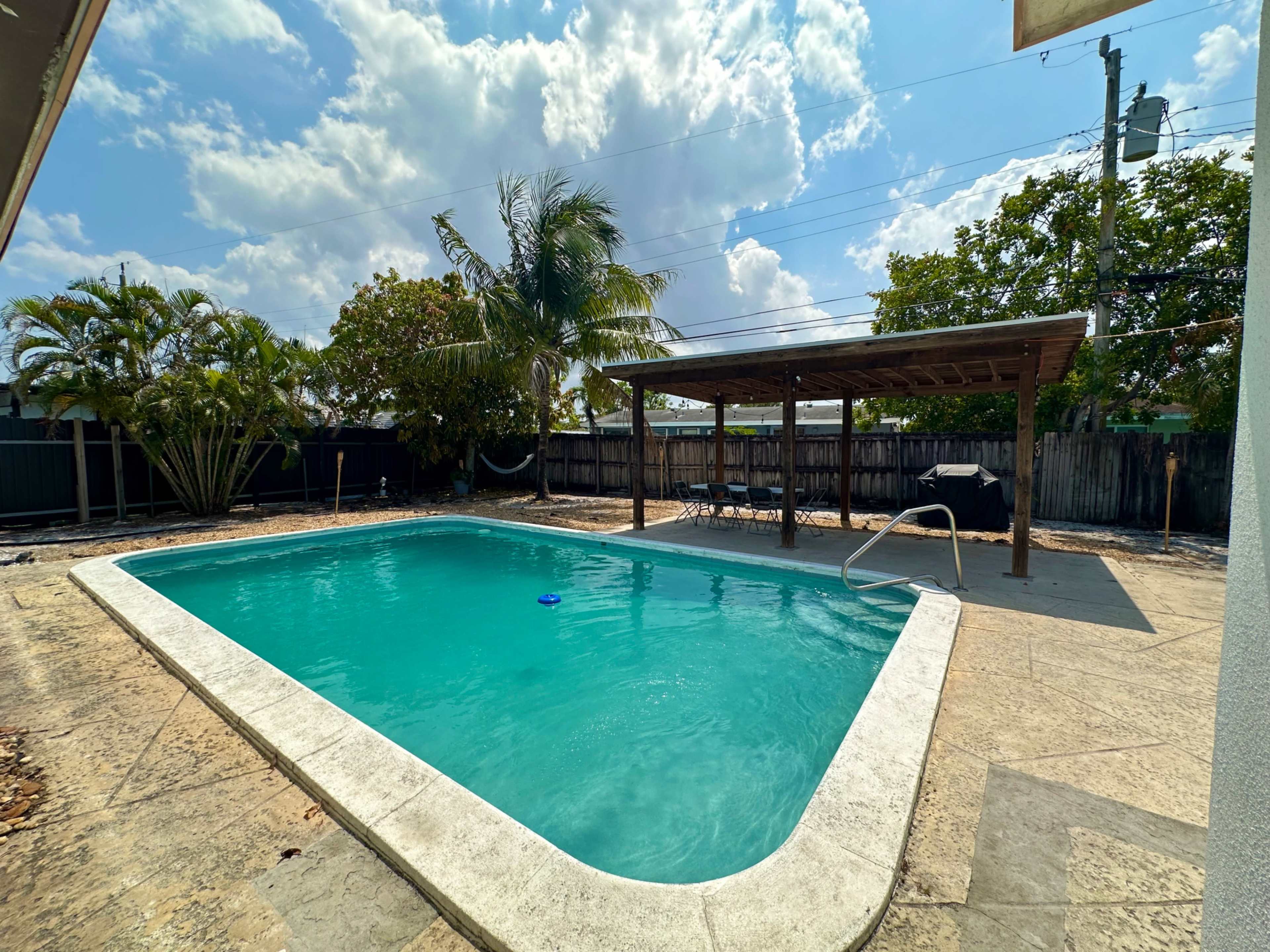 A rectangular swimming pool surrounded by concrete, with palm trees and a wooden structure in the background under a partly cloudy sky.
