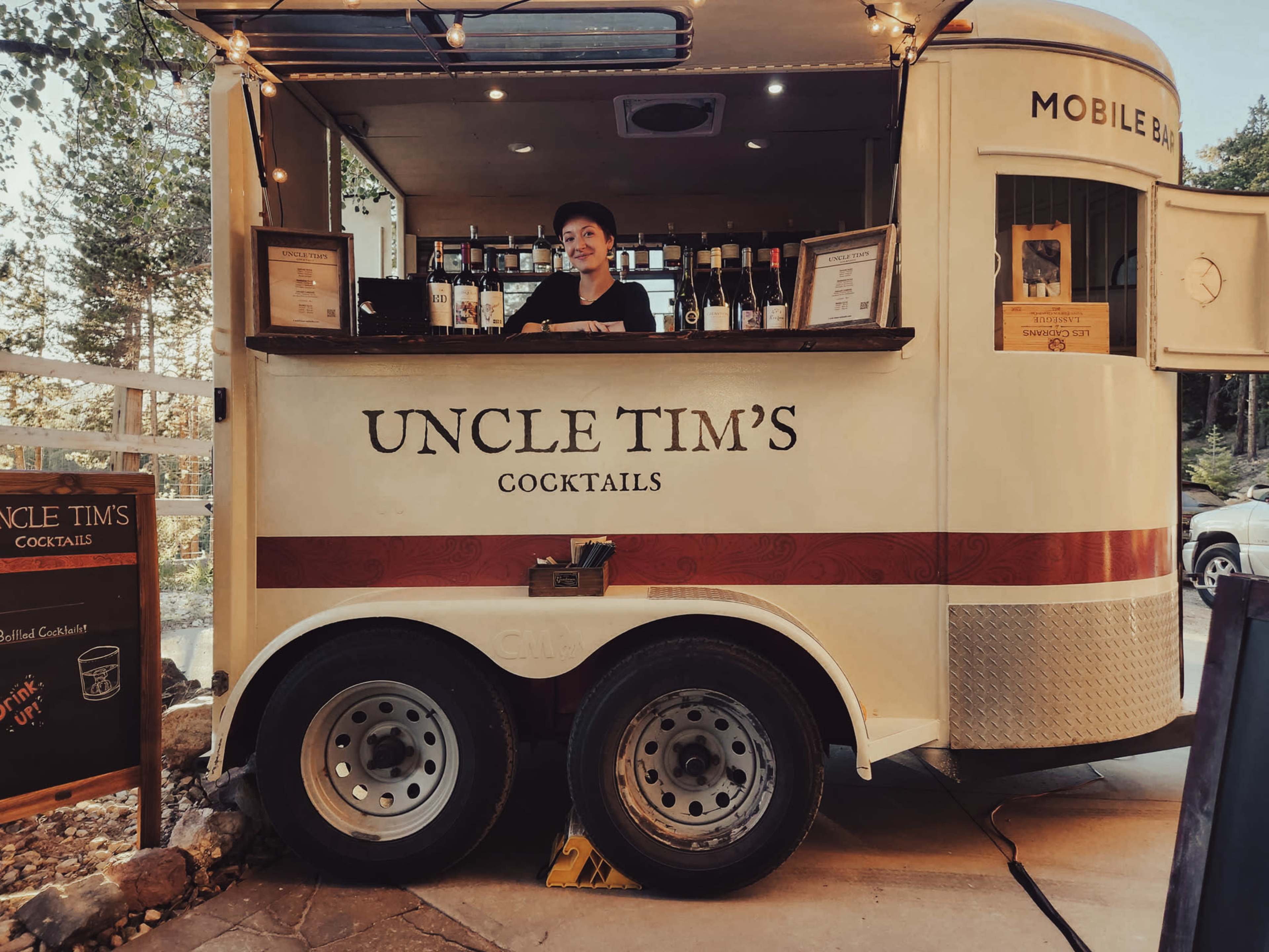 A mobile bar named "Uncle Tim's Cocktails" is set up with a smiling bartender behind it, surrounded by various bottles and serving options.