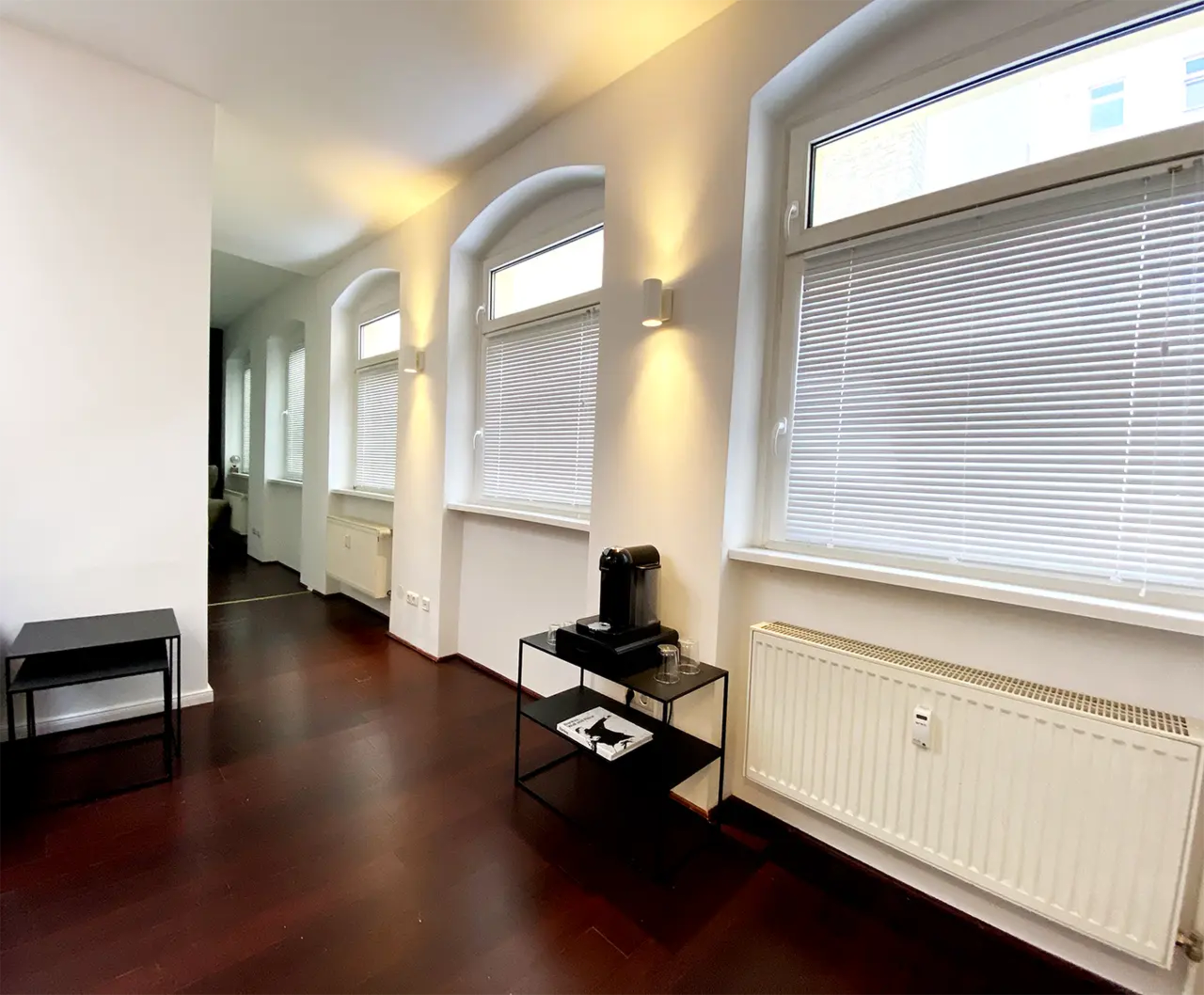 The image shows a brightly lit room with large windows, dark wooden flooring, and a black table holding a coffee maker and a few items.