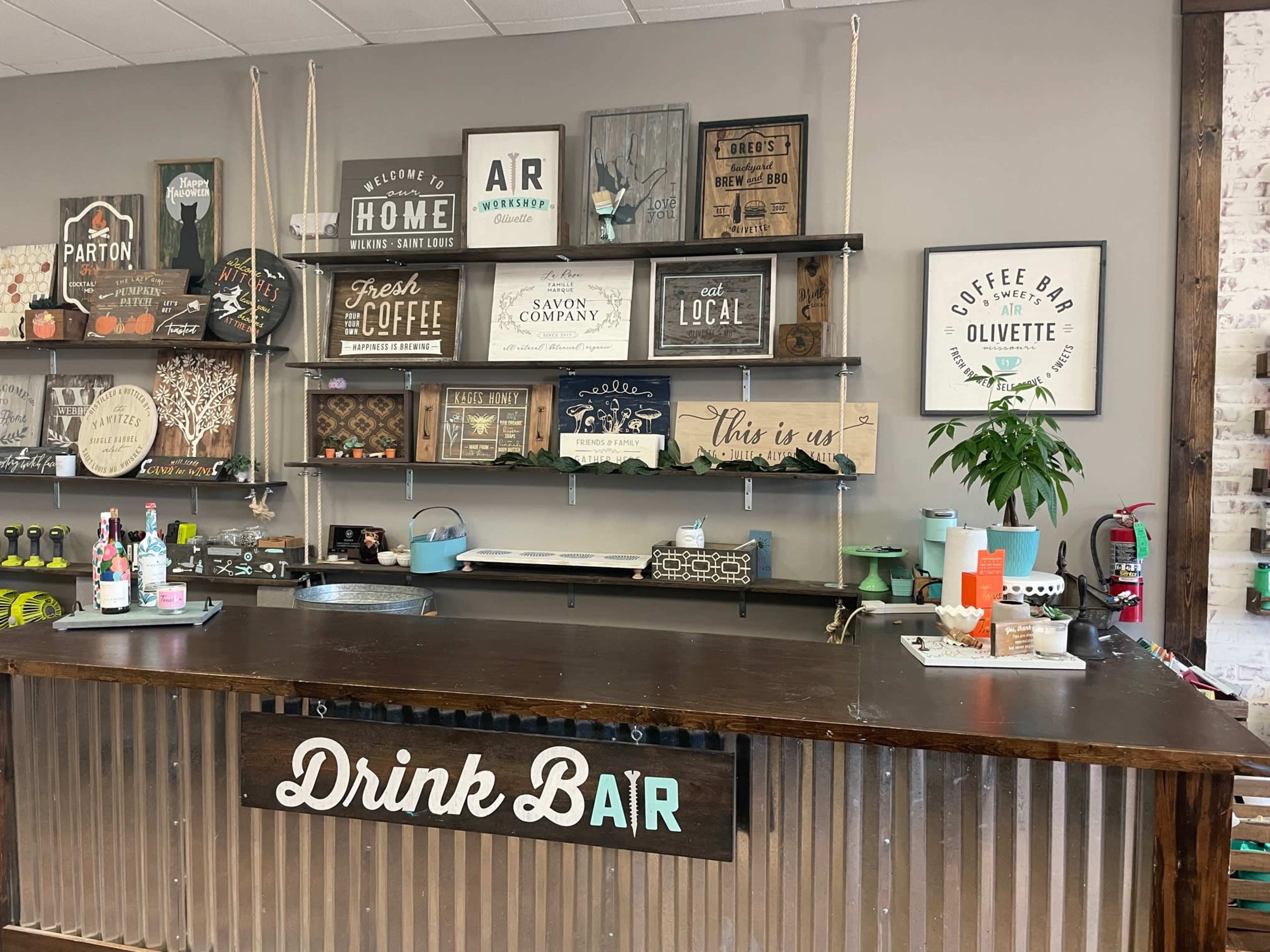 The image shows a drink bar with a wooden counter and shelves displaying various decorative signs and plants on a gray wall.
