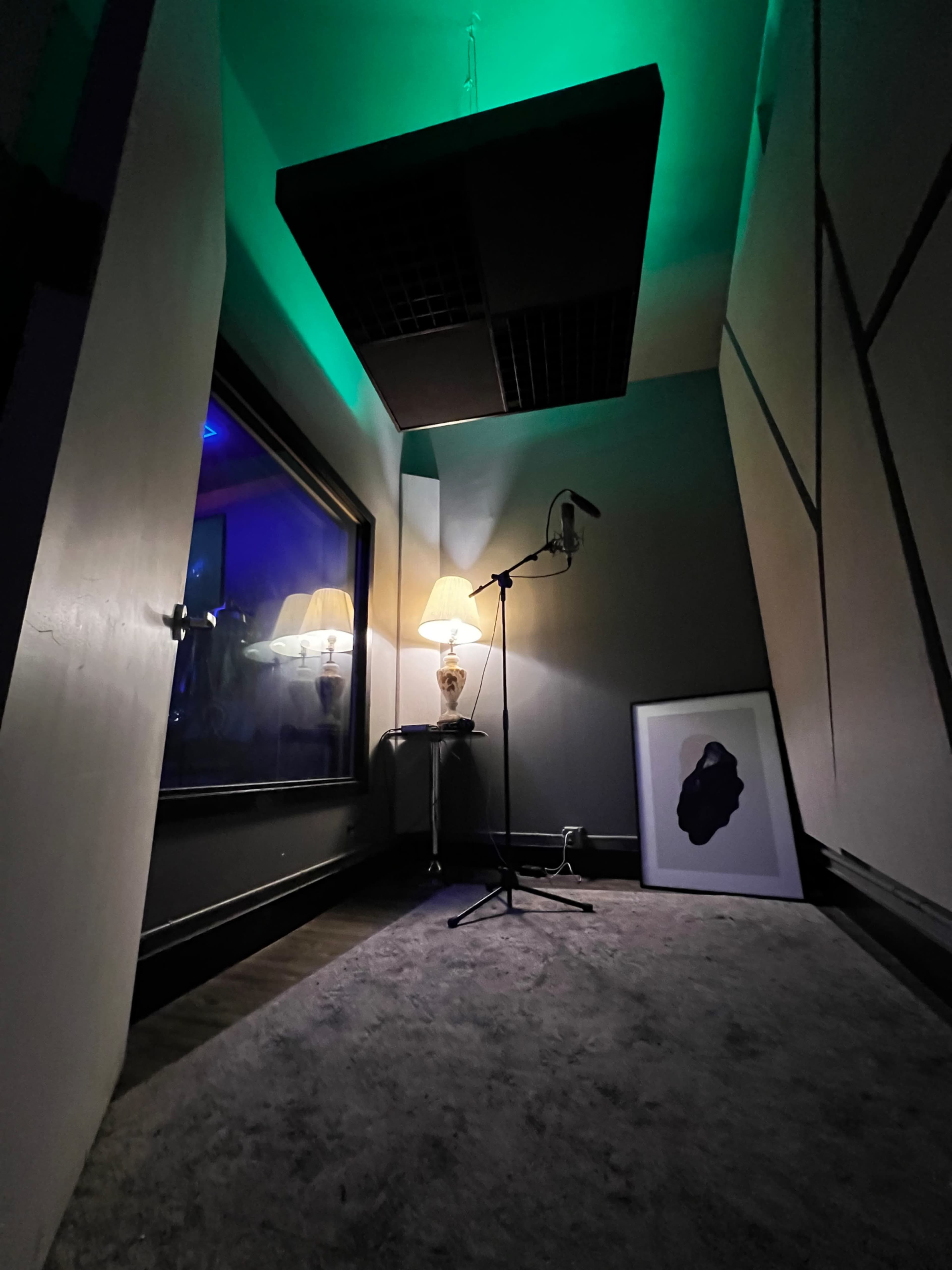 A dimly lit recording studio corner with a microphone, a lamp, a framed artwork, and illuminated green lighting.