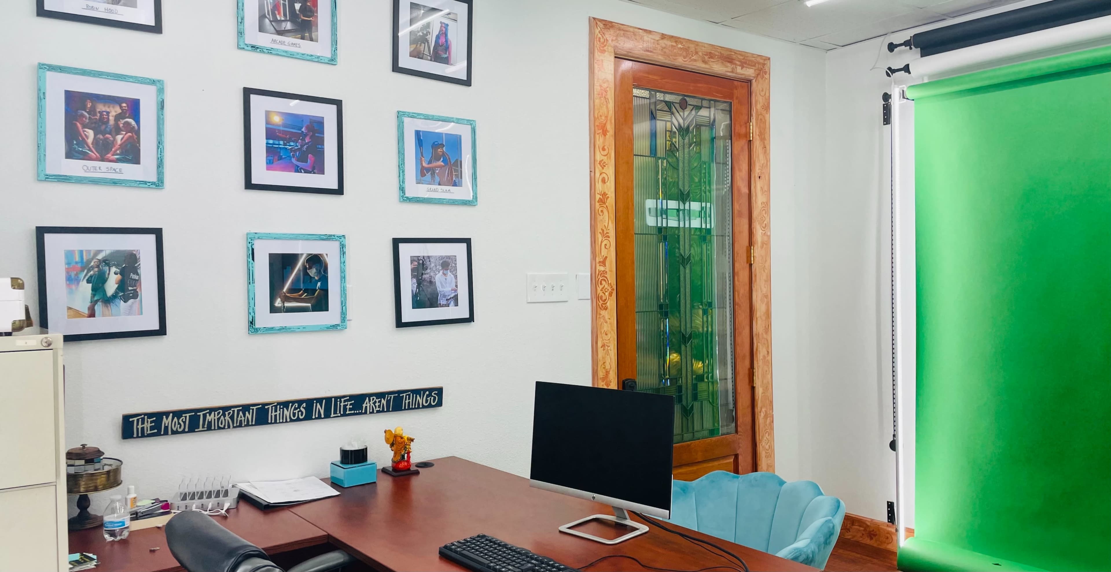An office space with a wooden desk, a computer setup, a wall decorated with framed photos, and a glass door next to a green backdrop.