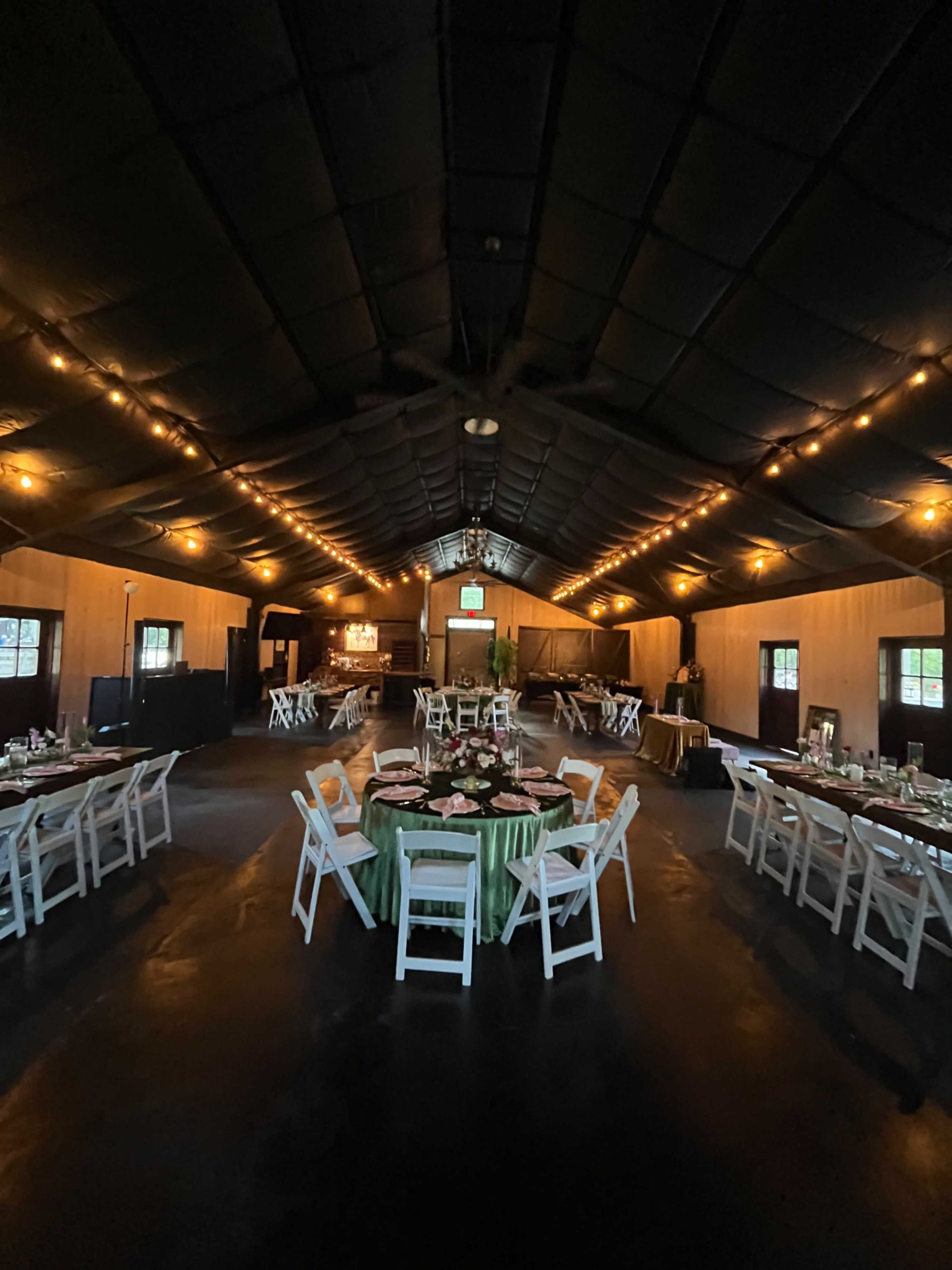 The image shows a spacious indoor venue with tables set for a gathering, illuminated by warm string lights along the ceiling.