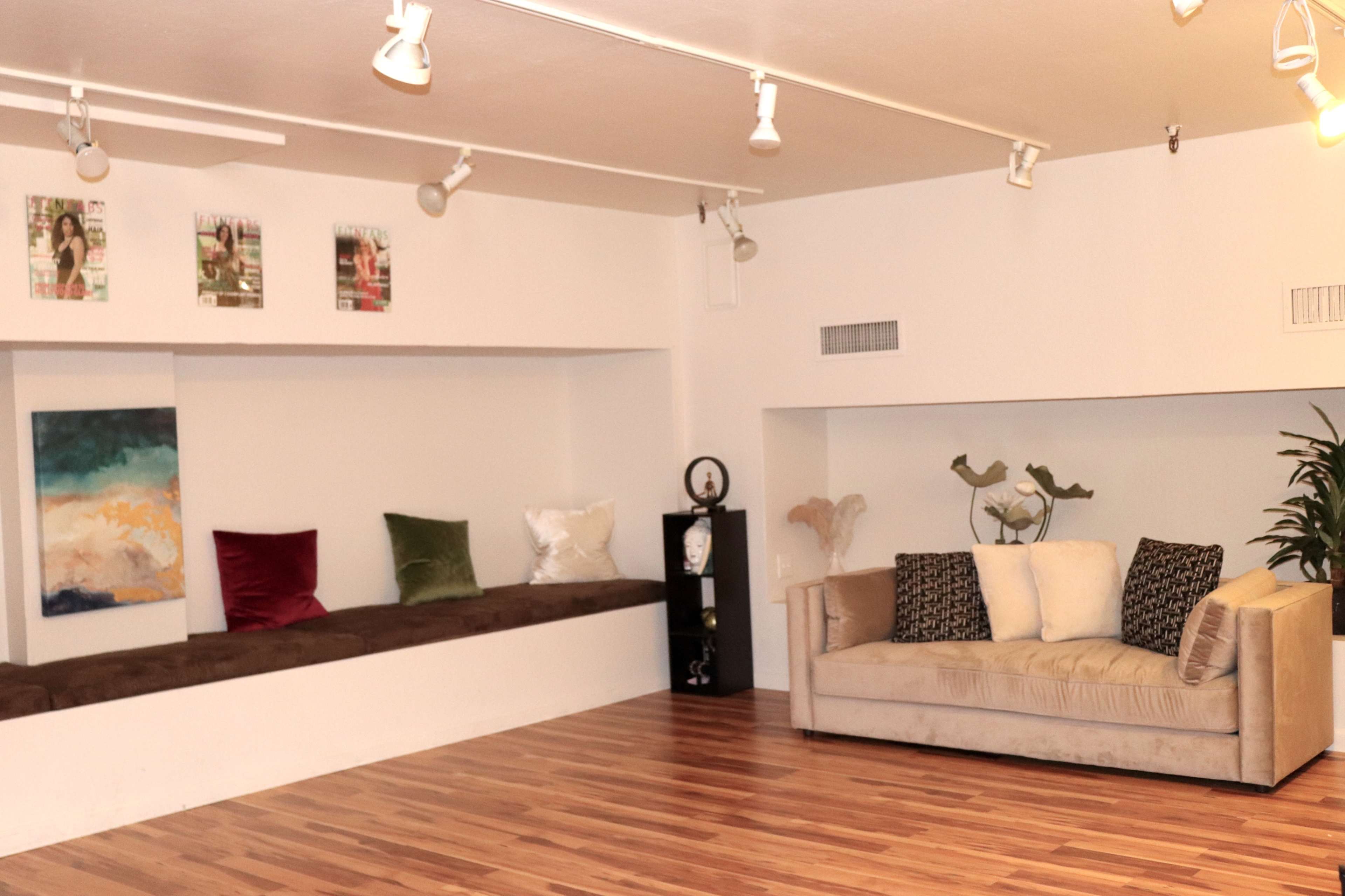 A spacious room with wooden flooring, featuring a long seating area with colorful cushions and a beige couch against the wall.