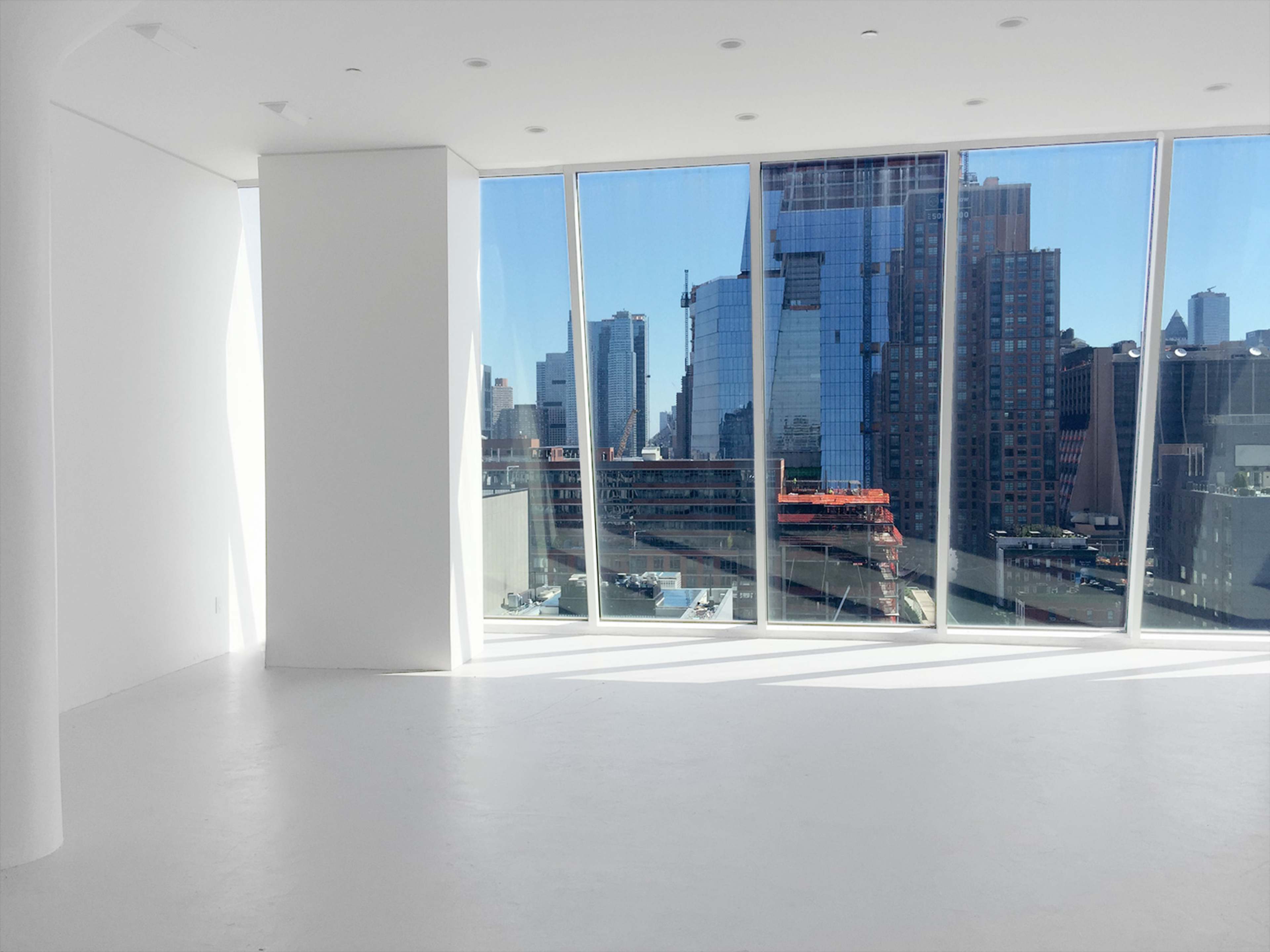 A spacious, minimalist room features large windows overlooking a city skyline with tall buildings and construction sites.