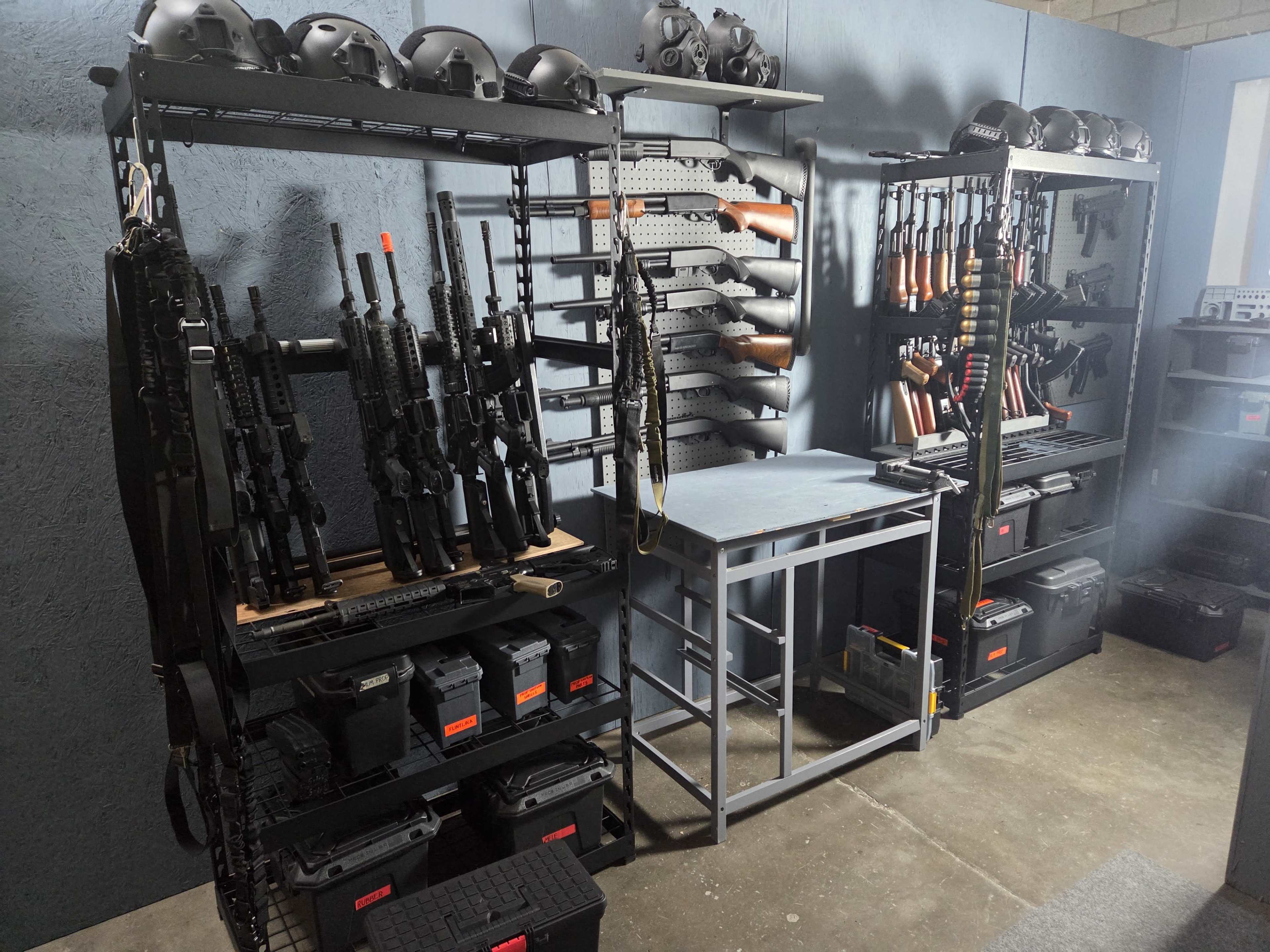 The image shows a room with two tightly organized racks holding various firearms and helmets, alongside a table and storage containers on the floor.