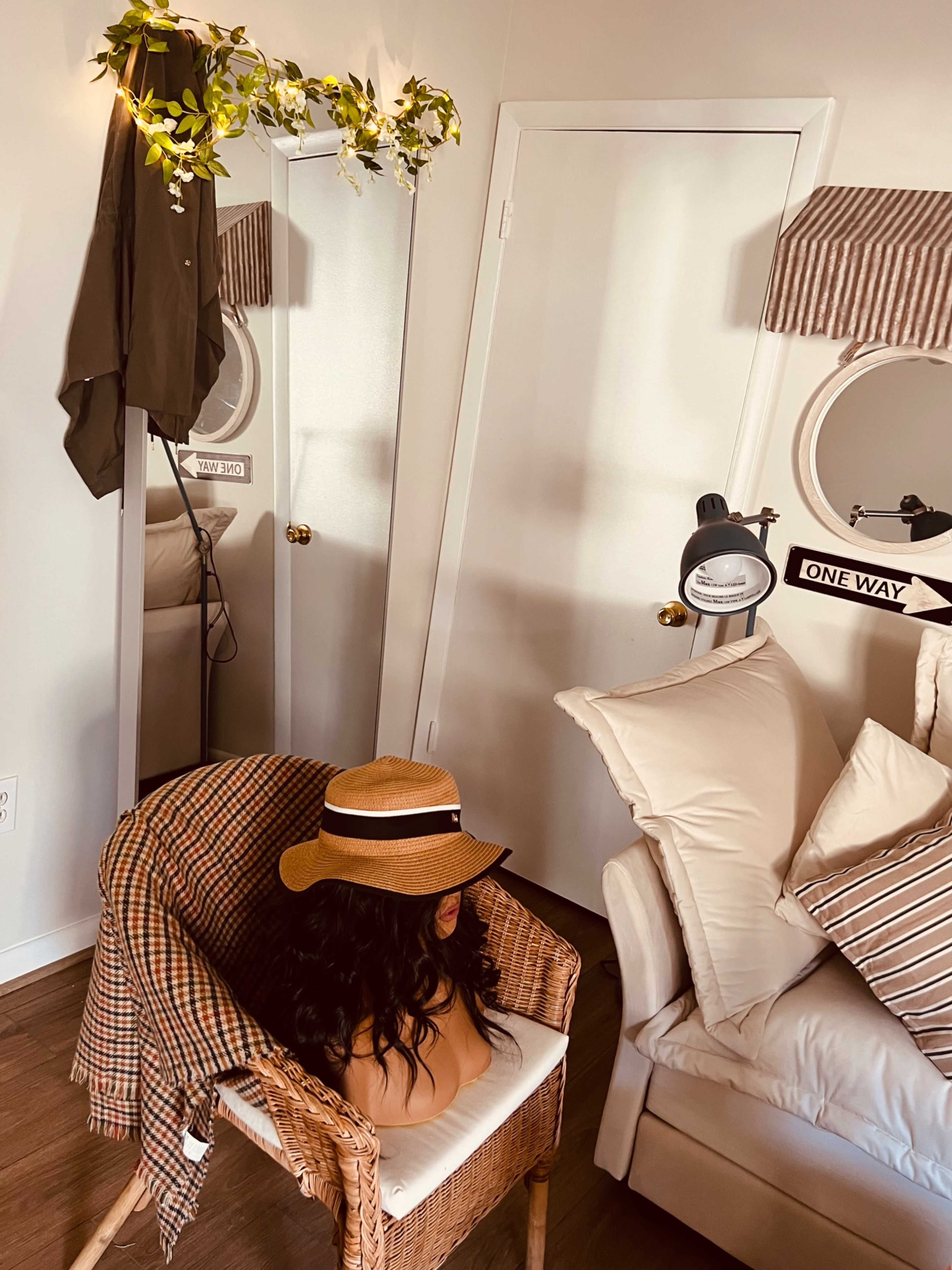 A hat sits on a wig mannequin placed in a chair next to a mirror in a softly lit room with neutral decor.