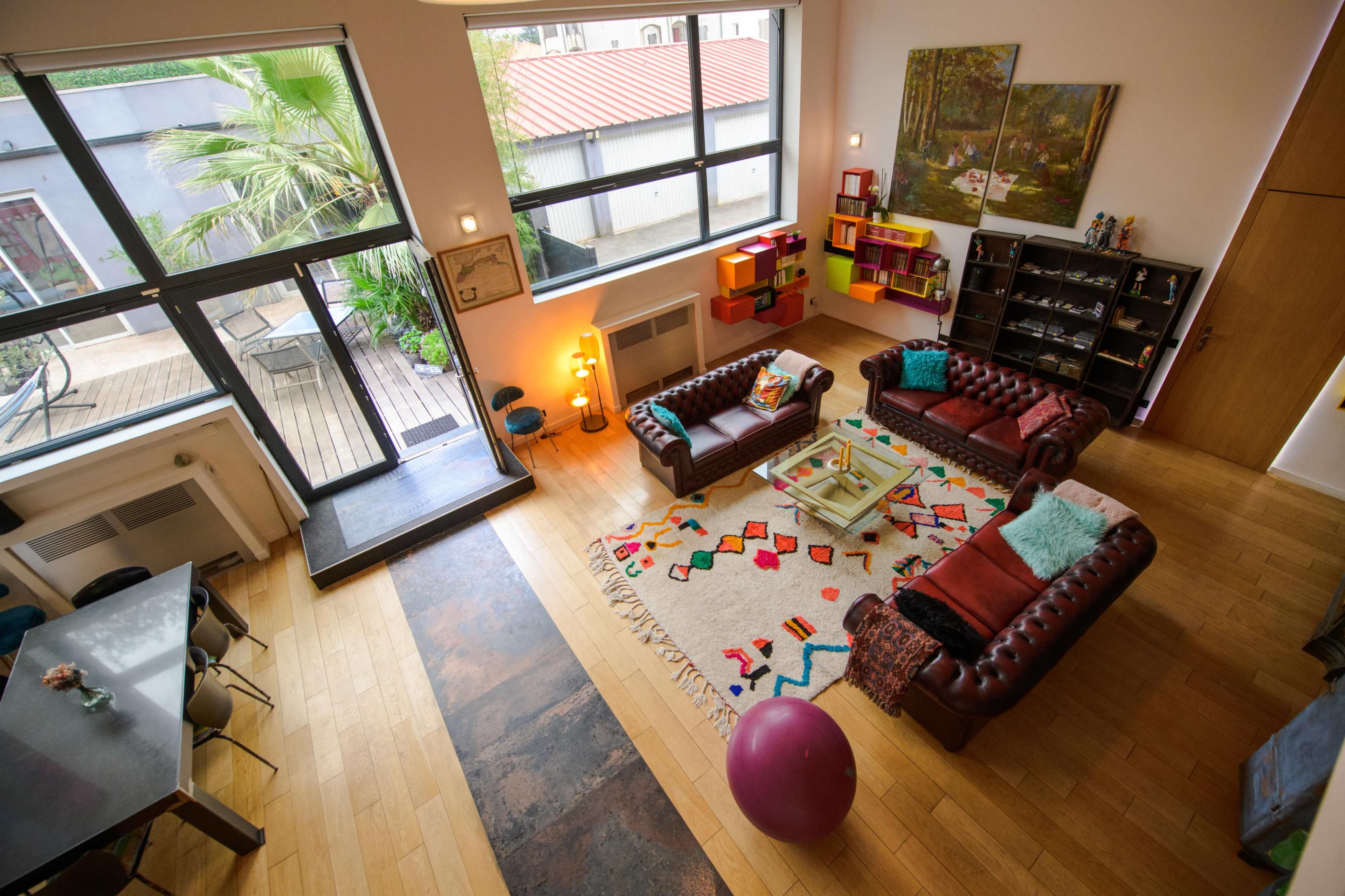 The image shows a spacious living area with two leather couches facing a colorful rug, surrounded by large windows that bring in natural light.