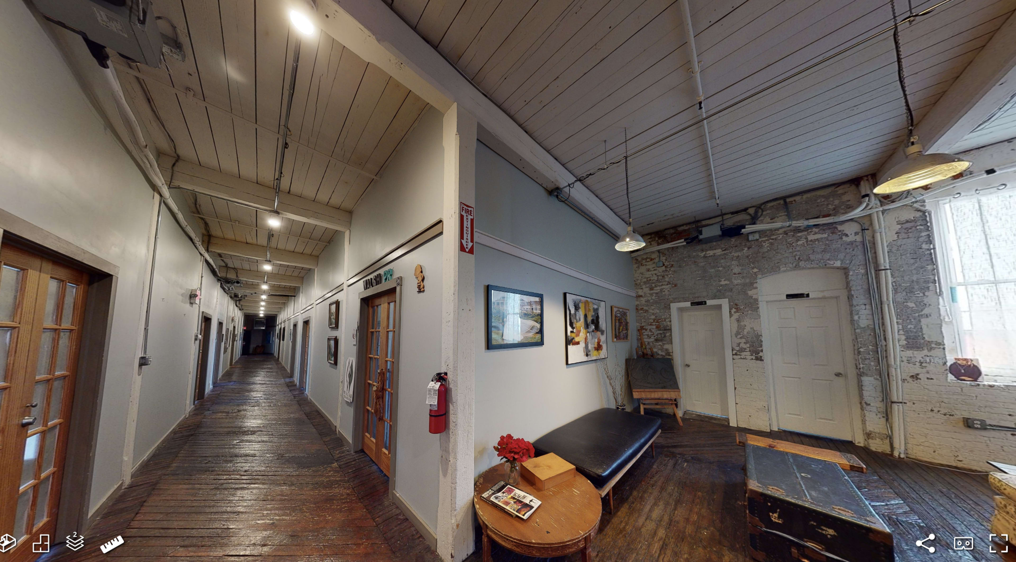 A long, narrow hallway with wooden floors, featuring several doorways along each side and a bench against one wall.