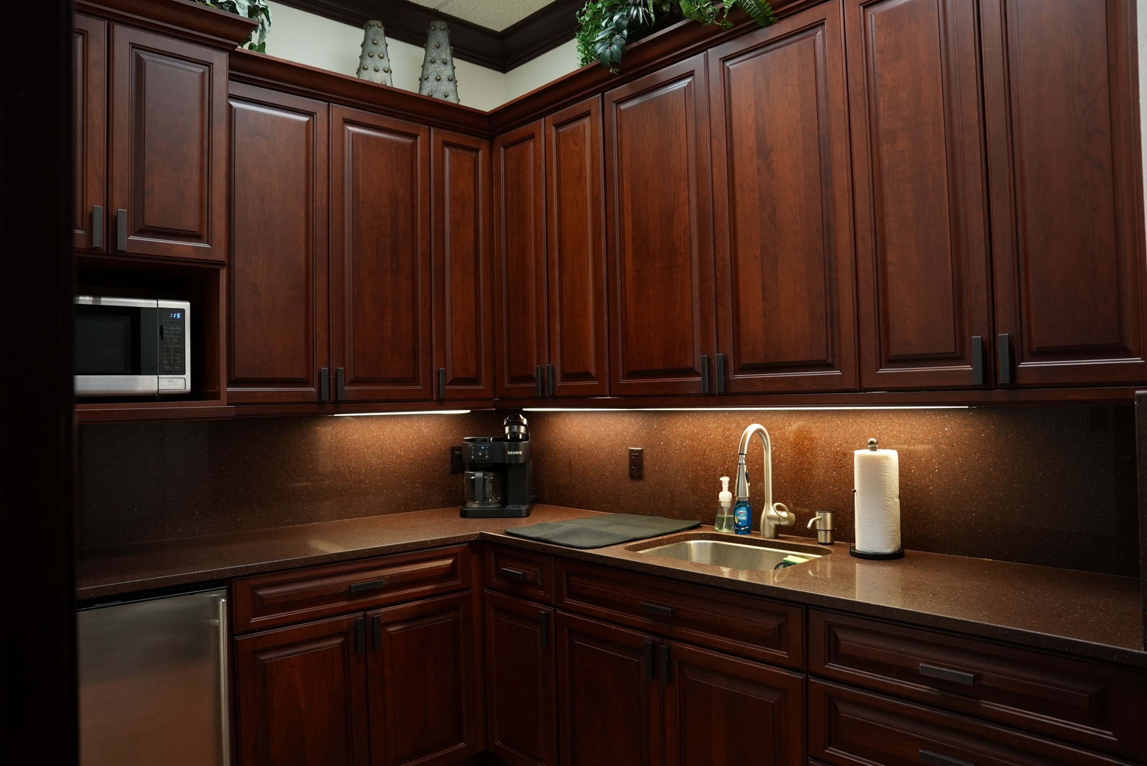 The image shows a kitchen with dark wood cabinetry, a stainless steel dishwasher, and a countertop featuring a sink, coffee maker, and paper towel holder.