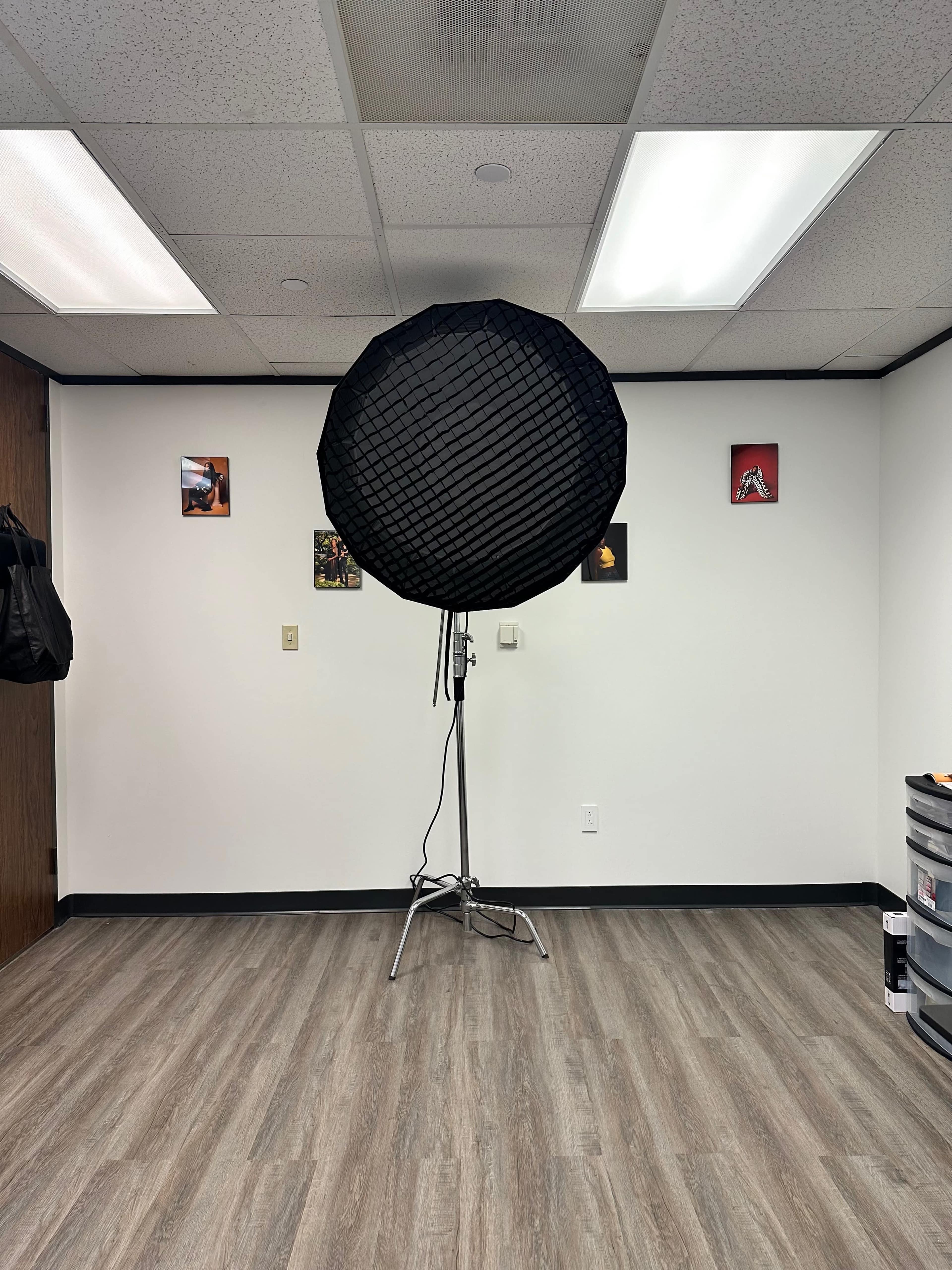 A large softbox light is positioned in the center of an empty room with gray walls and a wooden floor, accompanied by a few framed pictures on the walls.