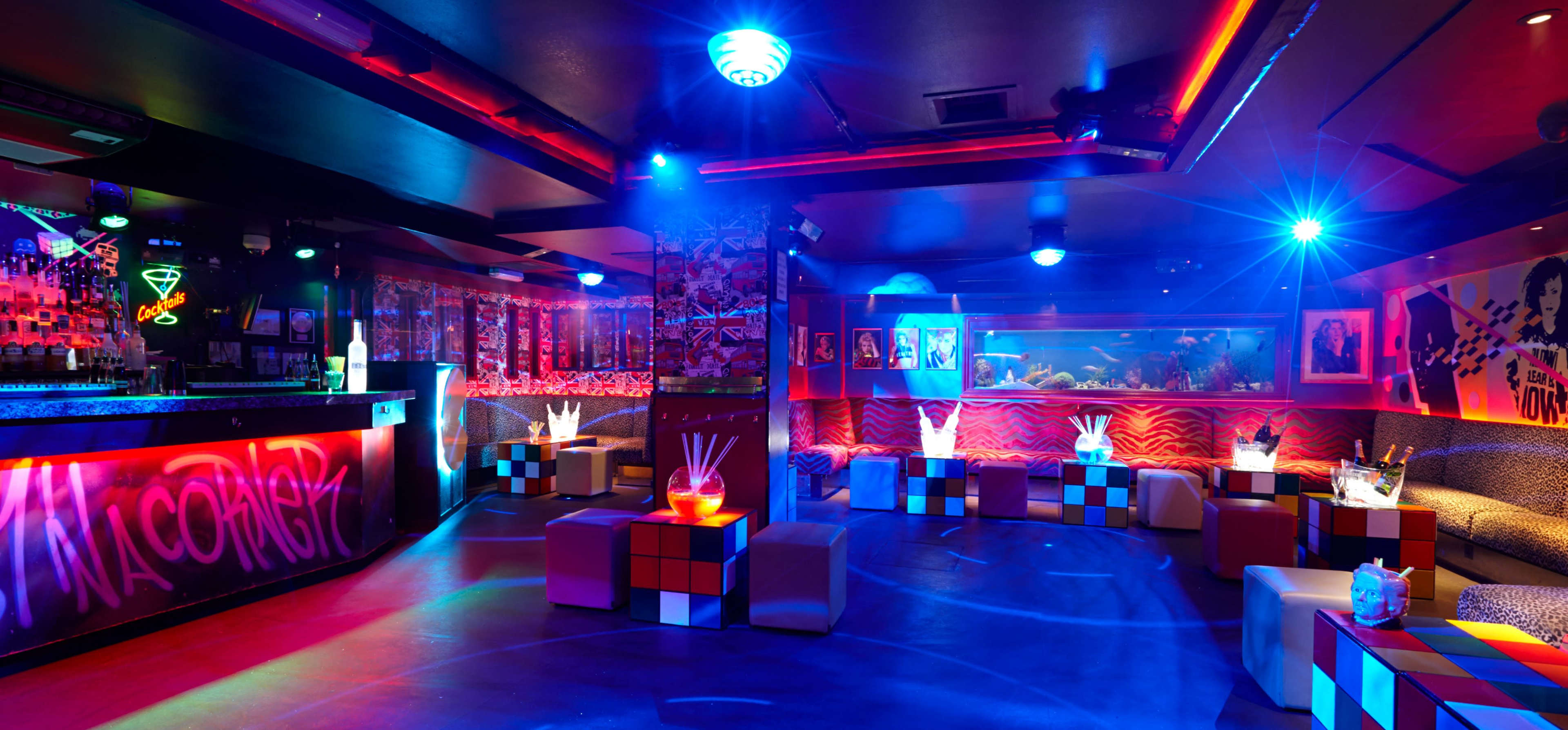 A vibrant nightclub features colorful lighting, a bar with illuminated drinks, and modern seating arranged around cube-shaped tables.