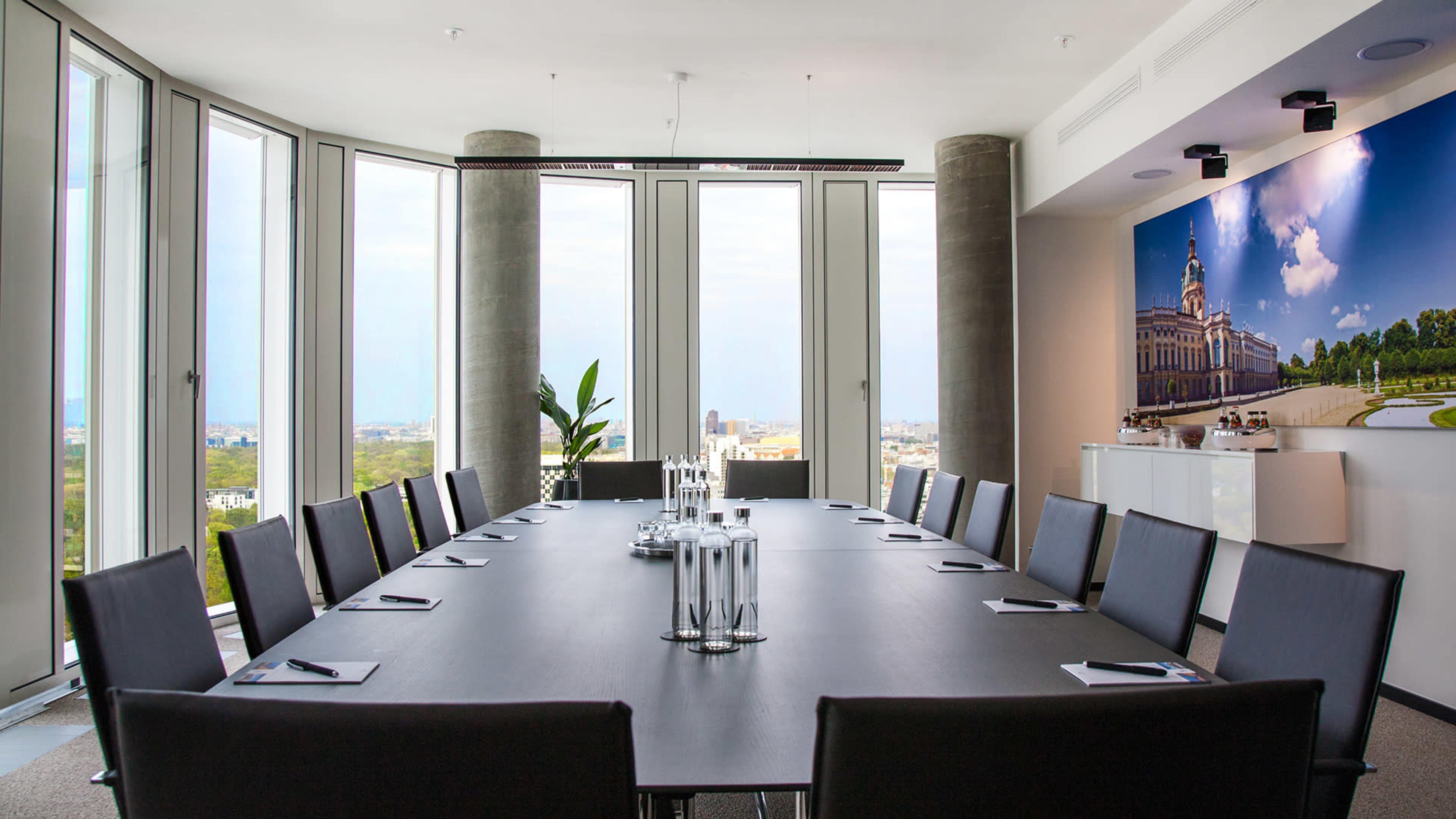 A modern conference room features a long rectangular table surrounded by black chairs, large windows offering a city view, and a wall display with a scenic image.