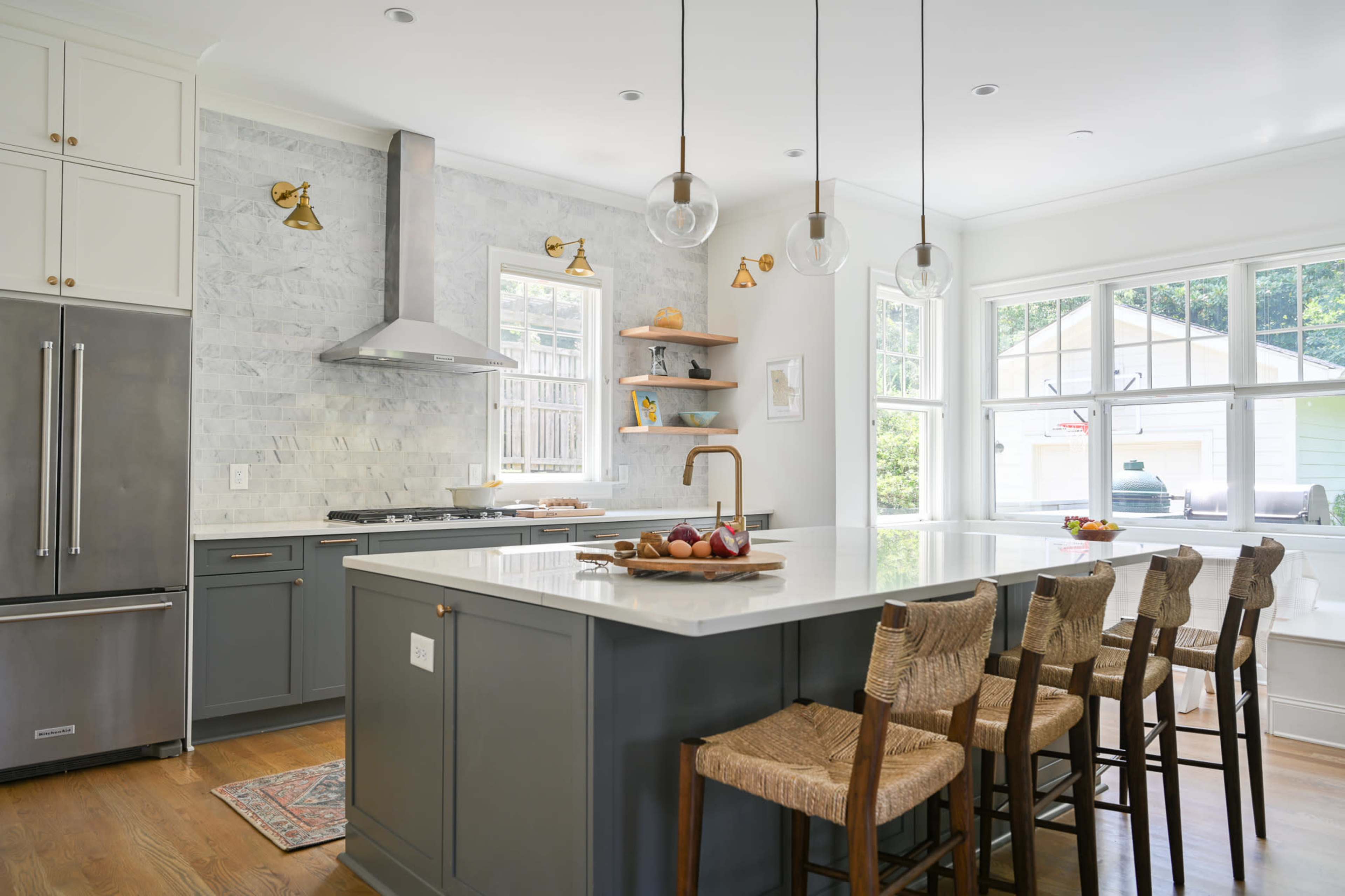 A modern kitchen features a large island with eight woven bar stools, marble countertops, stainless steel appliances, and a backsplash of light-colored tiles.