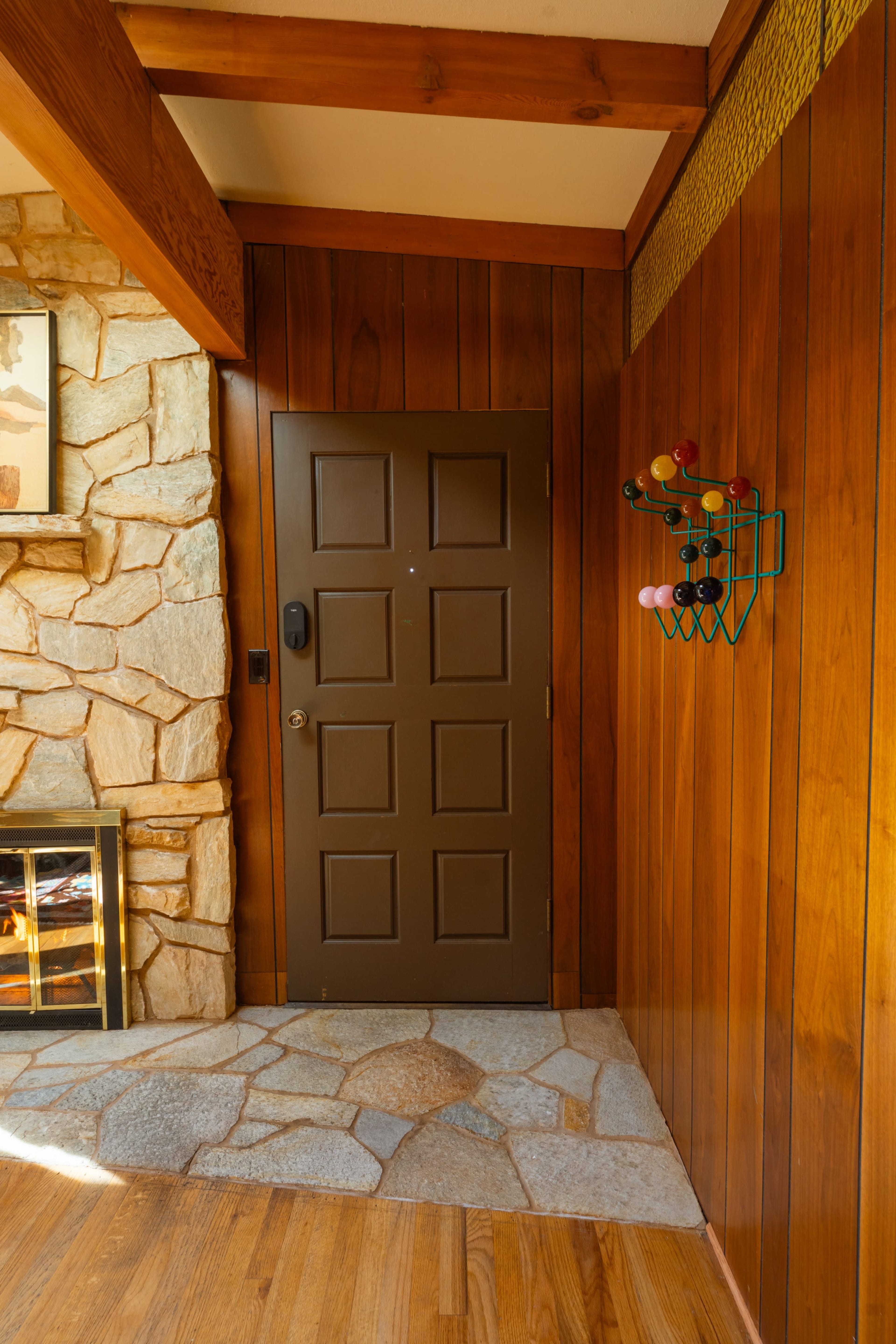 The image shows a wooden-paneled interior wall with a brown door, a stone fireplace, and a colorful wall-mounted wine rack.