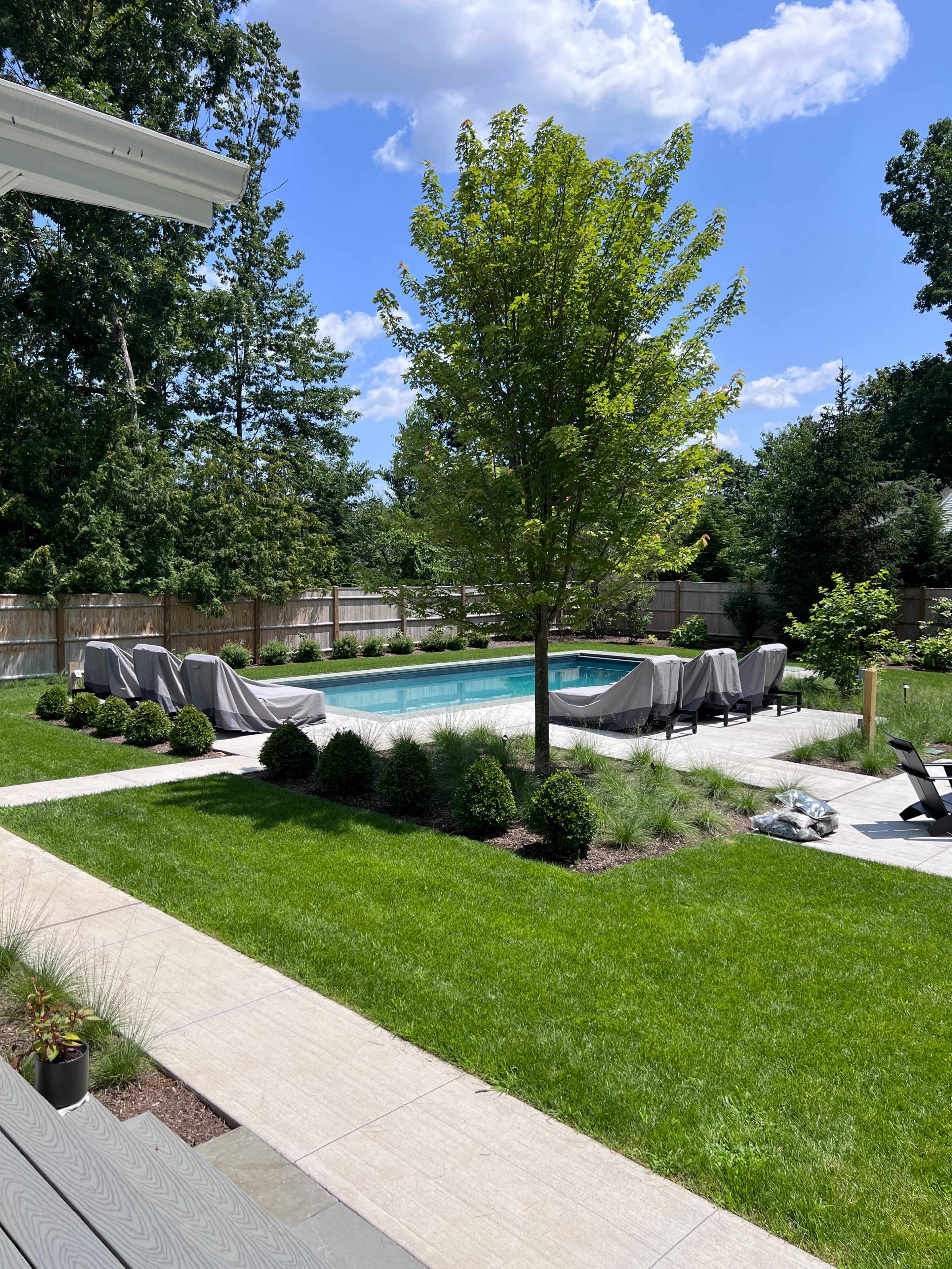 The image shows a landscaped backyard featuring a swimming pool surrounded by neatly trimmed grass, hedges, and shaded seating areas.