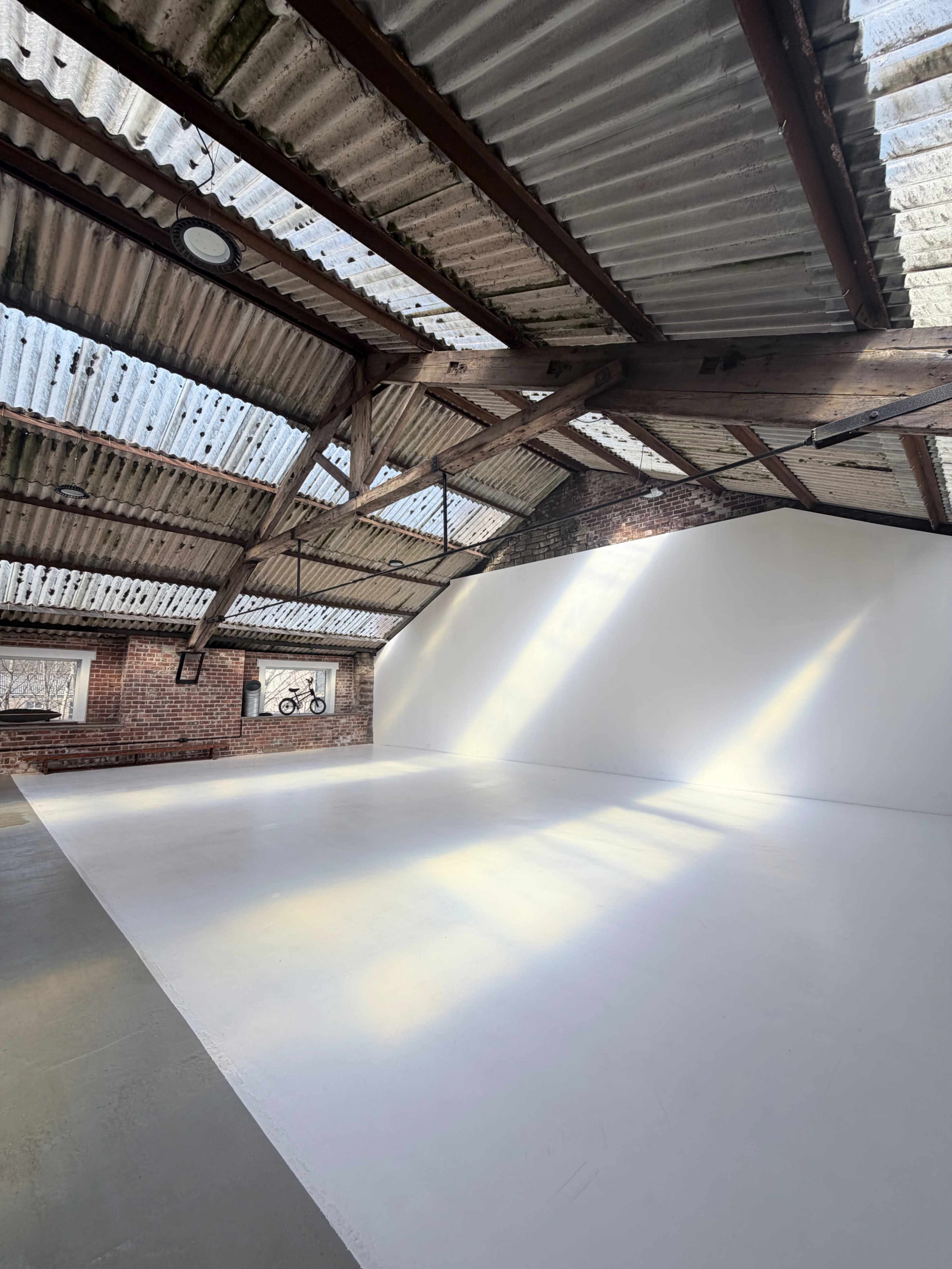 The image shows an industrial-style interior with a white wall, exposed brick, wooden beams, and skylights.