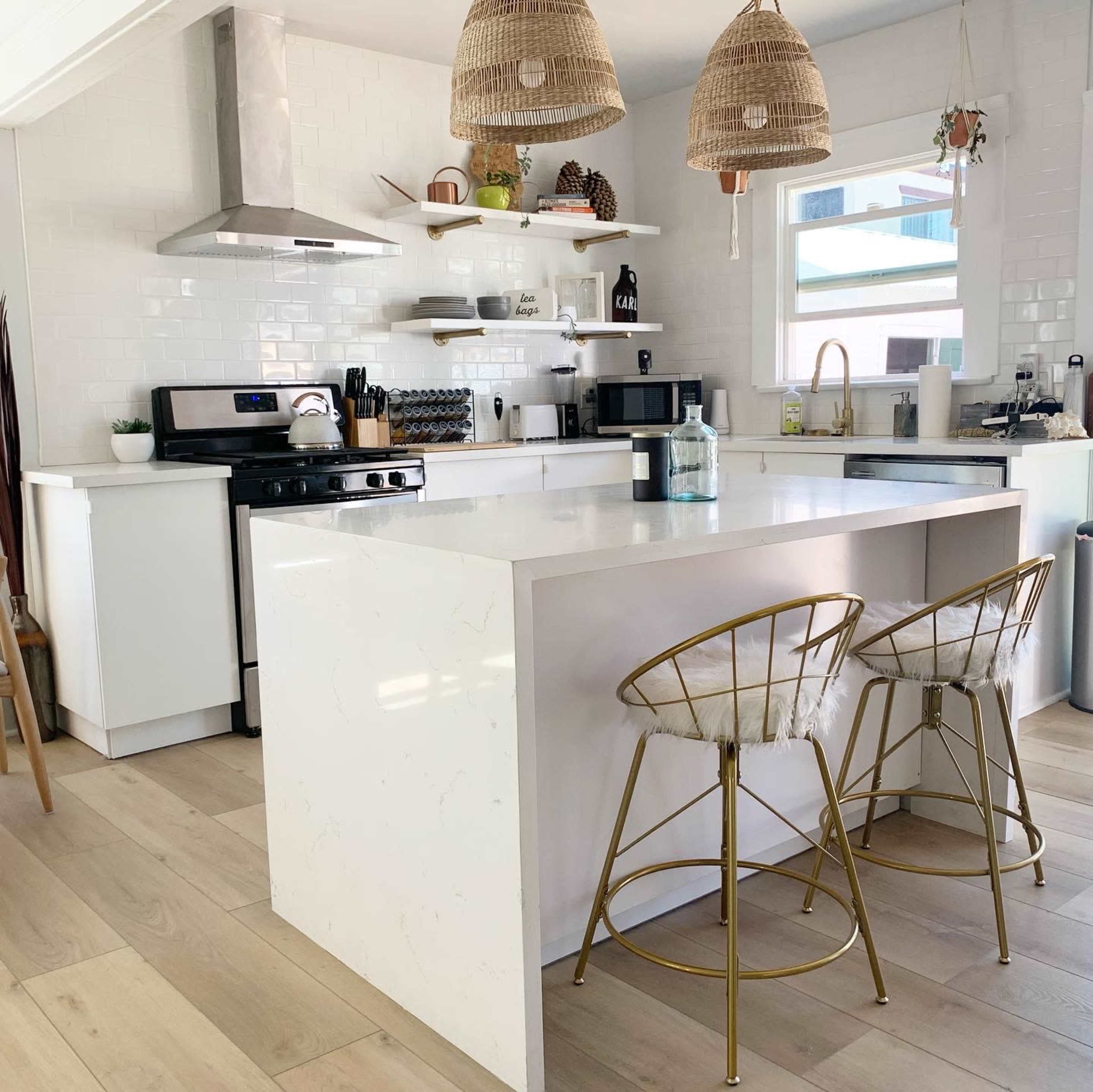 A modern kitchen features a white countertop island with two gold bar stools, white cabinetry, and open shelving against a backdrop of white tile.