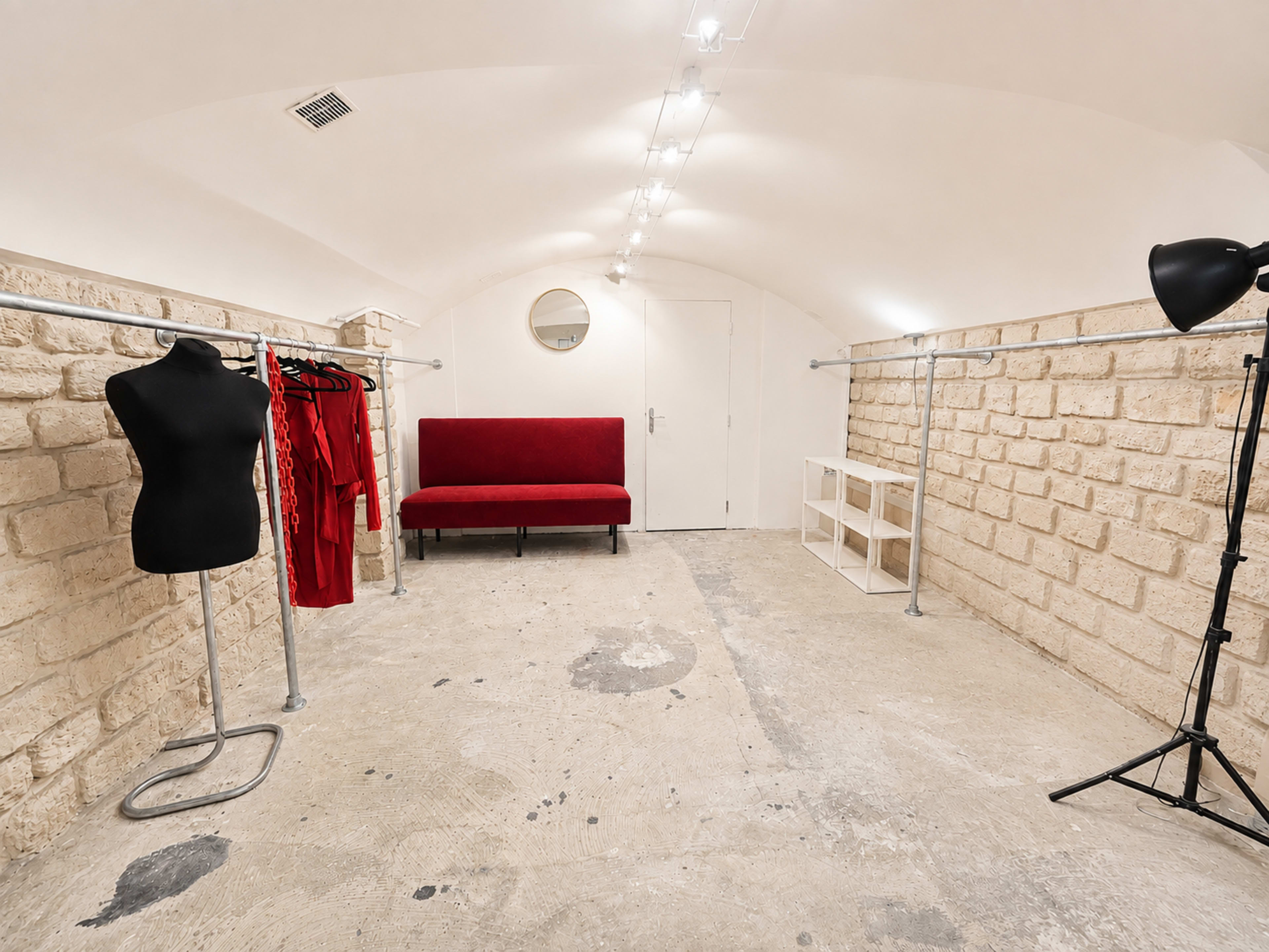 The image shows a minimalist dressing area with a red sofa, a clothing rack with red garments, a mirror on the wall, and exposed stone walls.